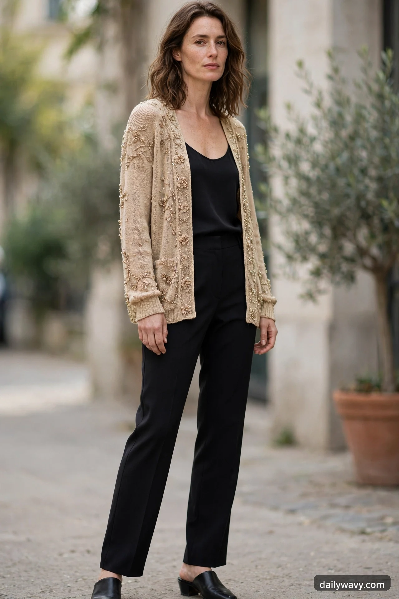 A woman stands on a city sidewalk wearing an open champagne beaded cardigan over a black camisole, black crepe trousers and black leather mules.