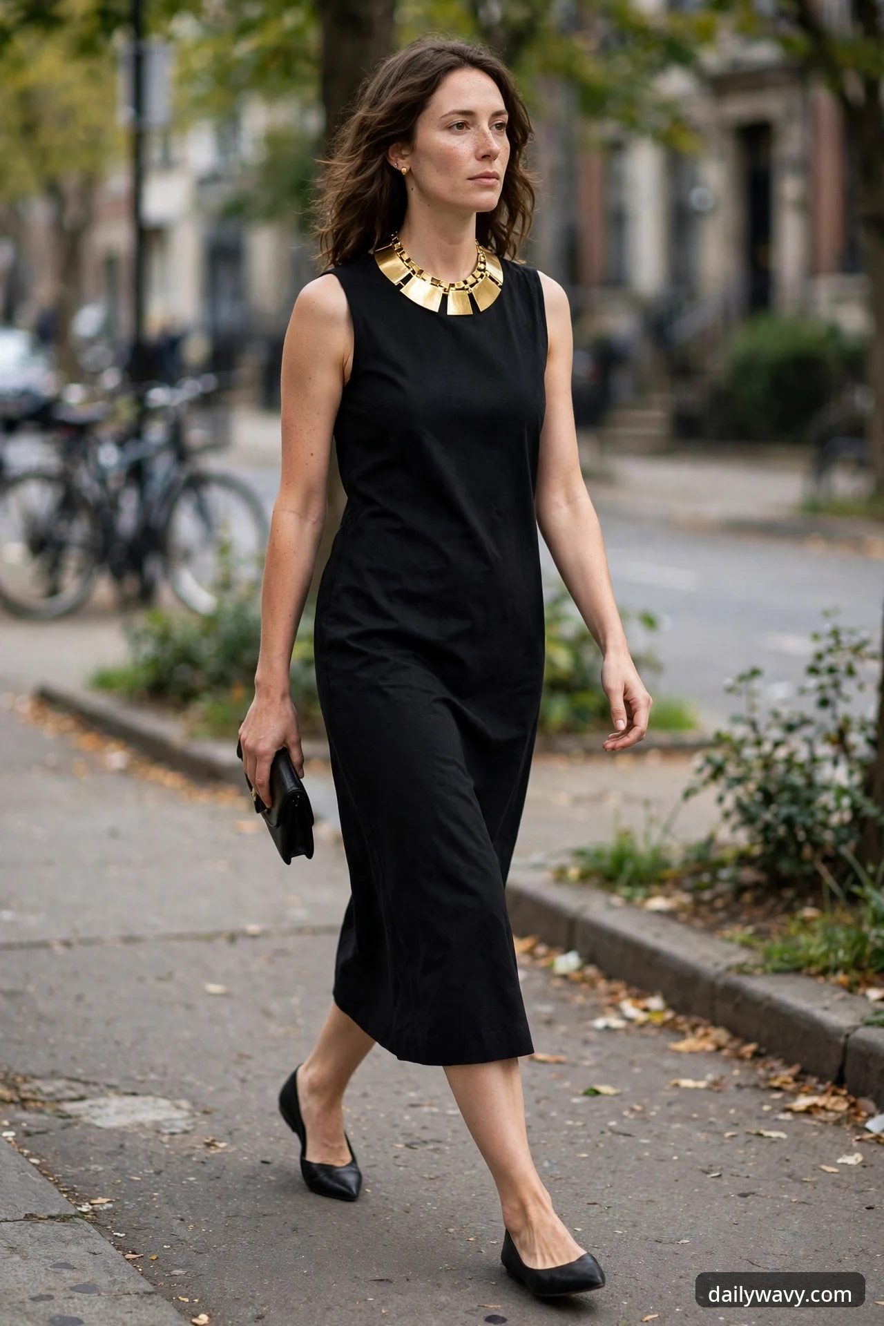 A woman walks down a city street wearing a black sleeveless midi dress, a bold gold geometric statement necklace, black pointed flats and a small black clutch.