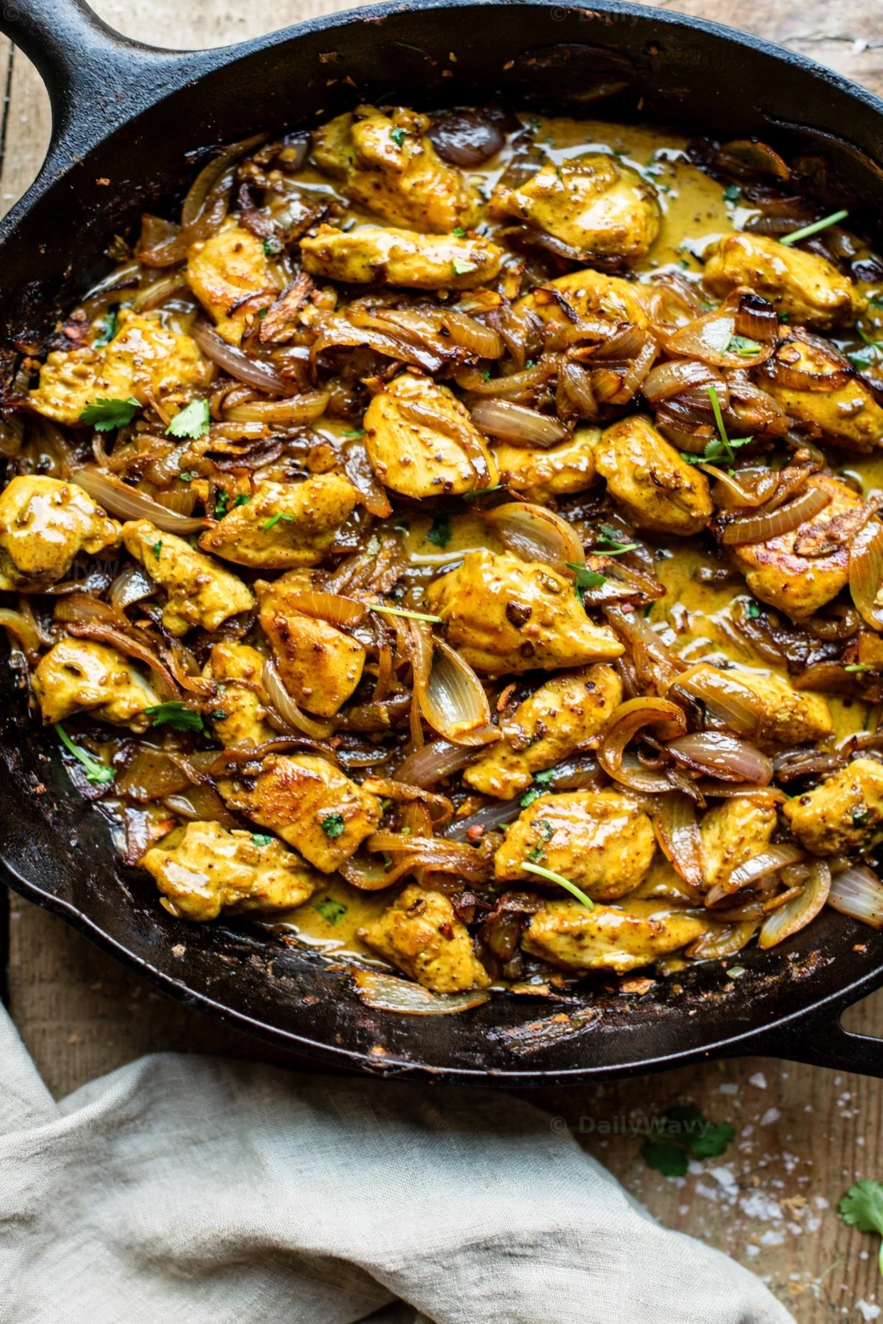 Chicken chunks and deeply caramelized onion slices fill a cast-iron skillet of coconut curry, seen from above with a linen napkin beside the pan.
