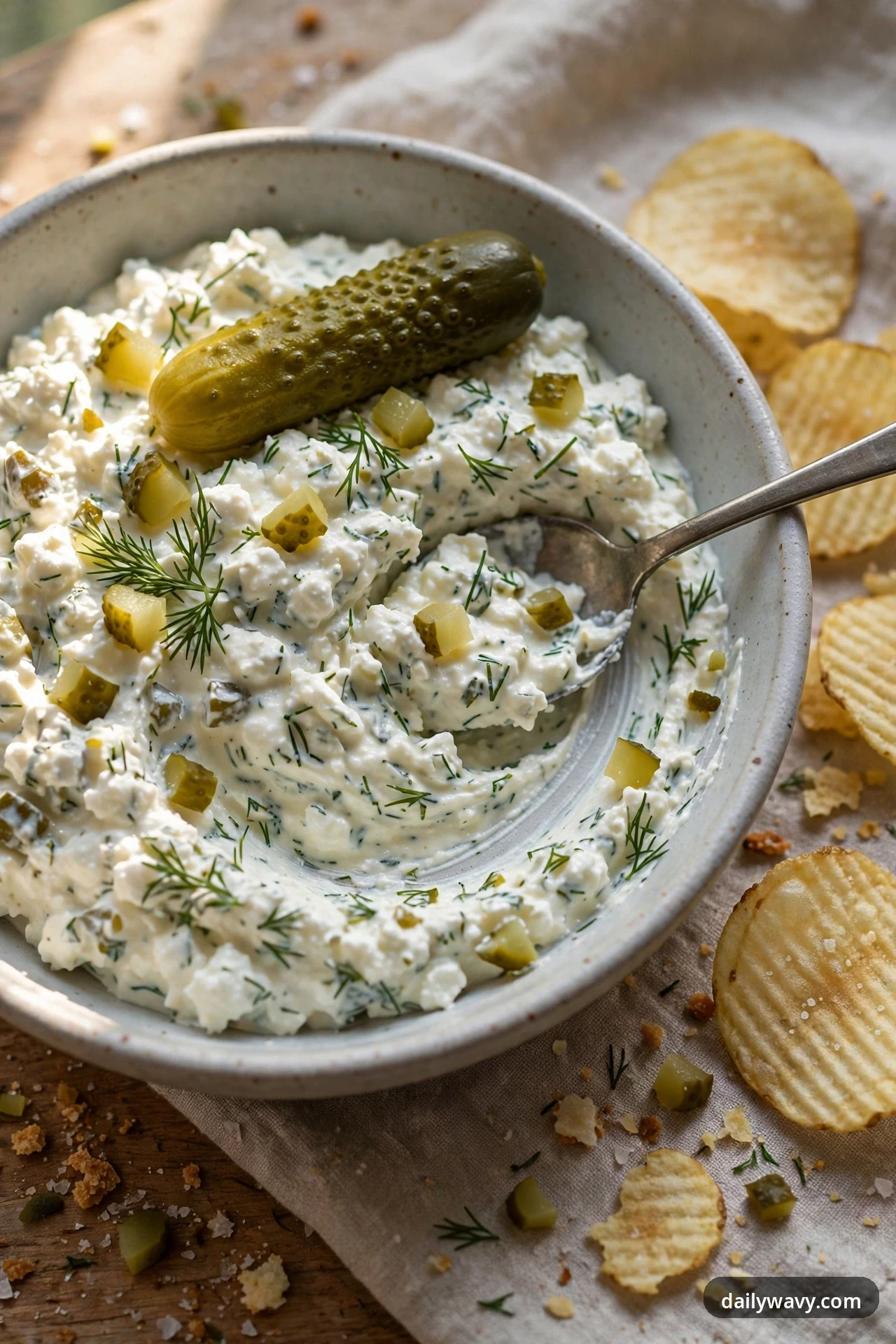 A bowl of cottage cheese dip is studded with chopped dill pickle and fresh dill, with a whole pickle laid across the top and ridged potato chips beside it.