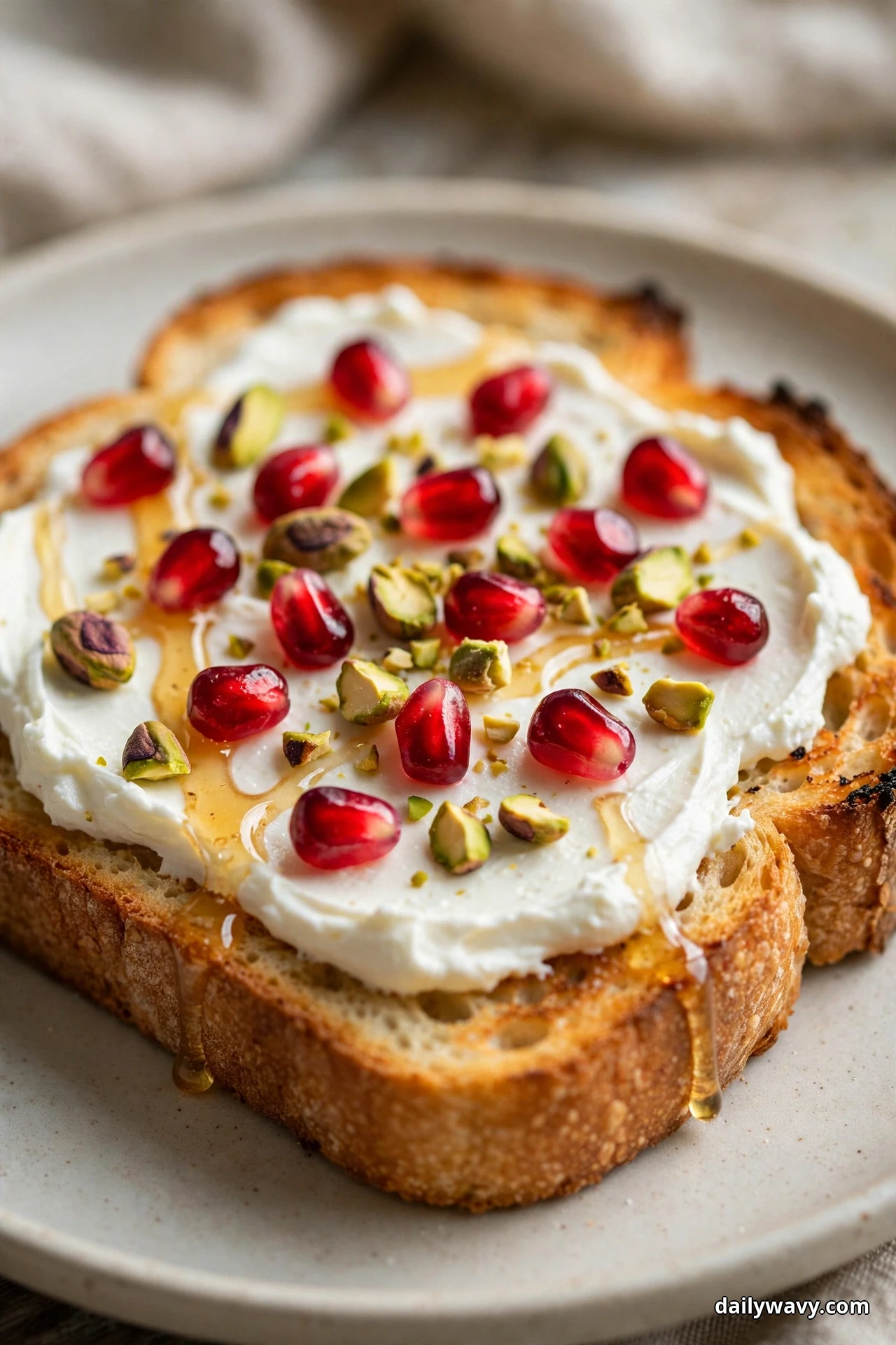 A slice of sourdough toast spread with cream cheese is scattered with ruby pomegranate seeds and chopped pistachios and finished with a honey drizzle