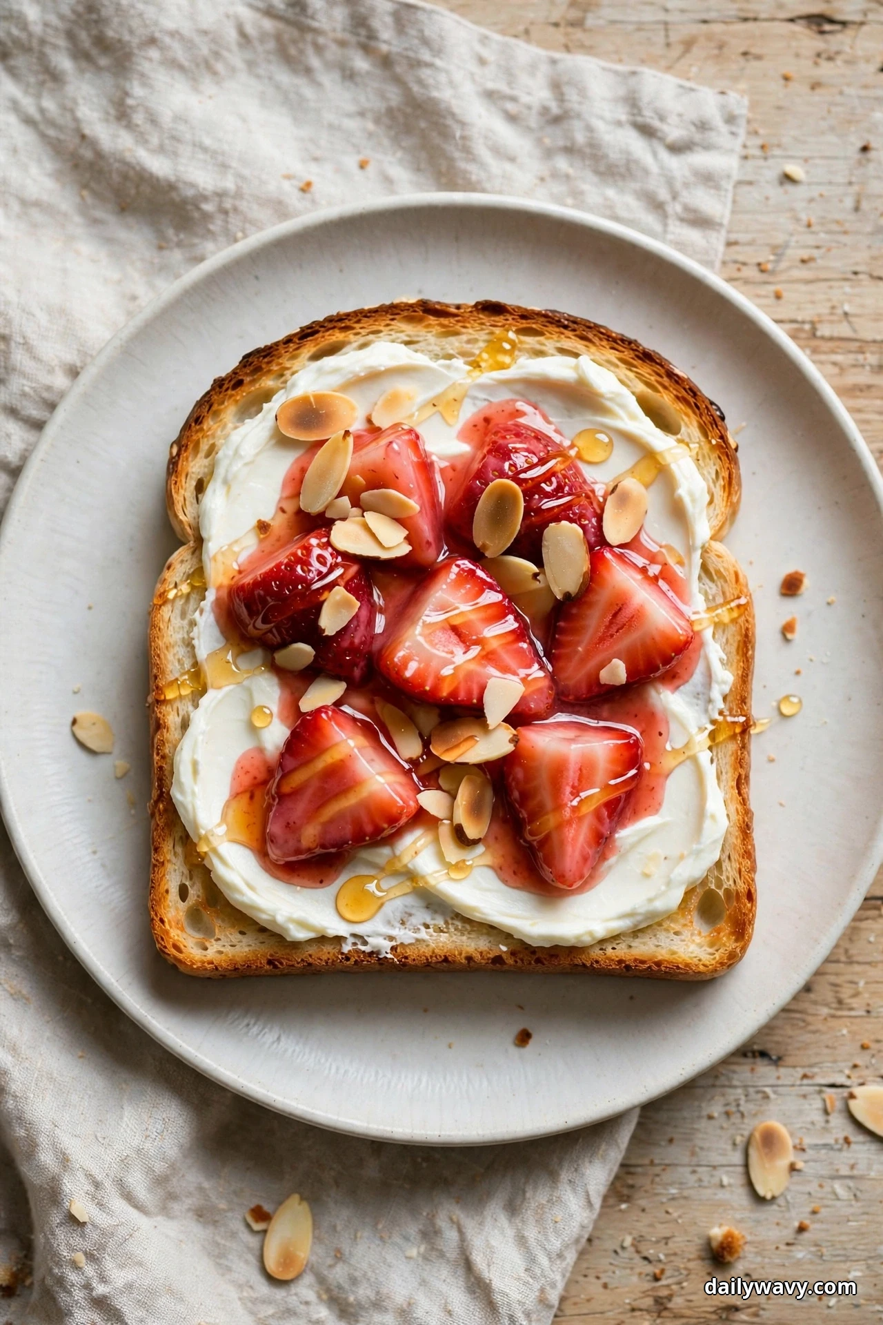 An overhead view shows a slice of toast spread with cream cheese and topped with glossy macerated strawberry quarters, toasted sliced almonds and a honey drizzle