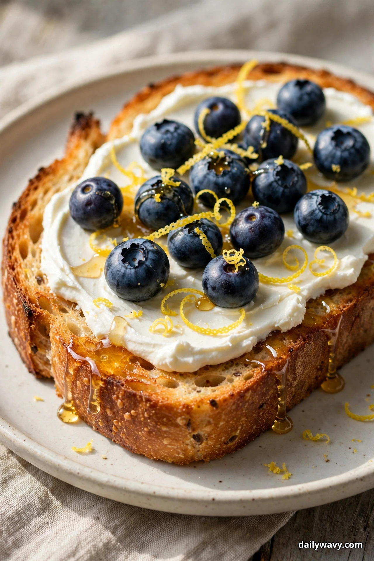 A slice of sourdough toast spread with cream cheese is topped with fresh blueberries and curled strips of lemon zest, with honey dripping over the crust