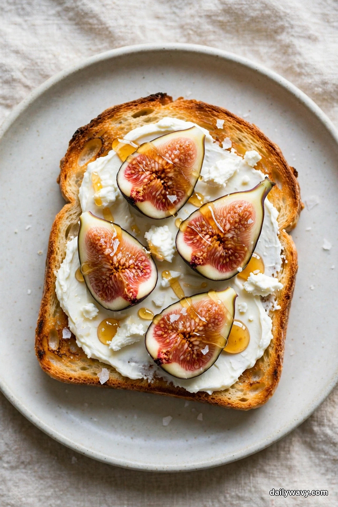 An overhead view shows a slice of sourdough toast spread with cream cheese and topped with fresh figs cut side up, crumbles of goat cheese, a honey drizzle and flaky sea salt