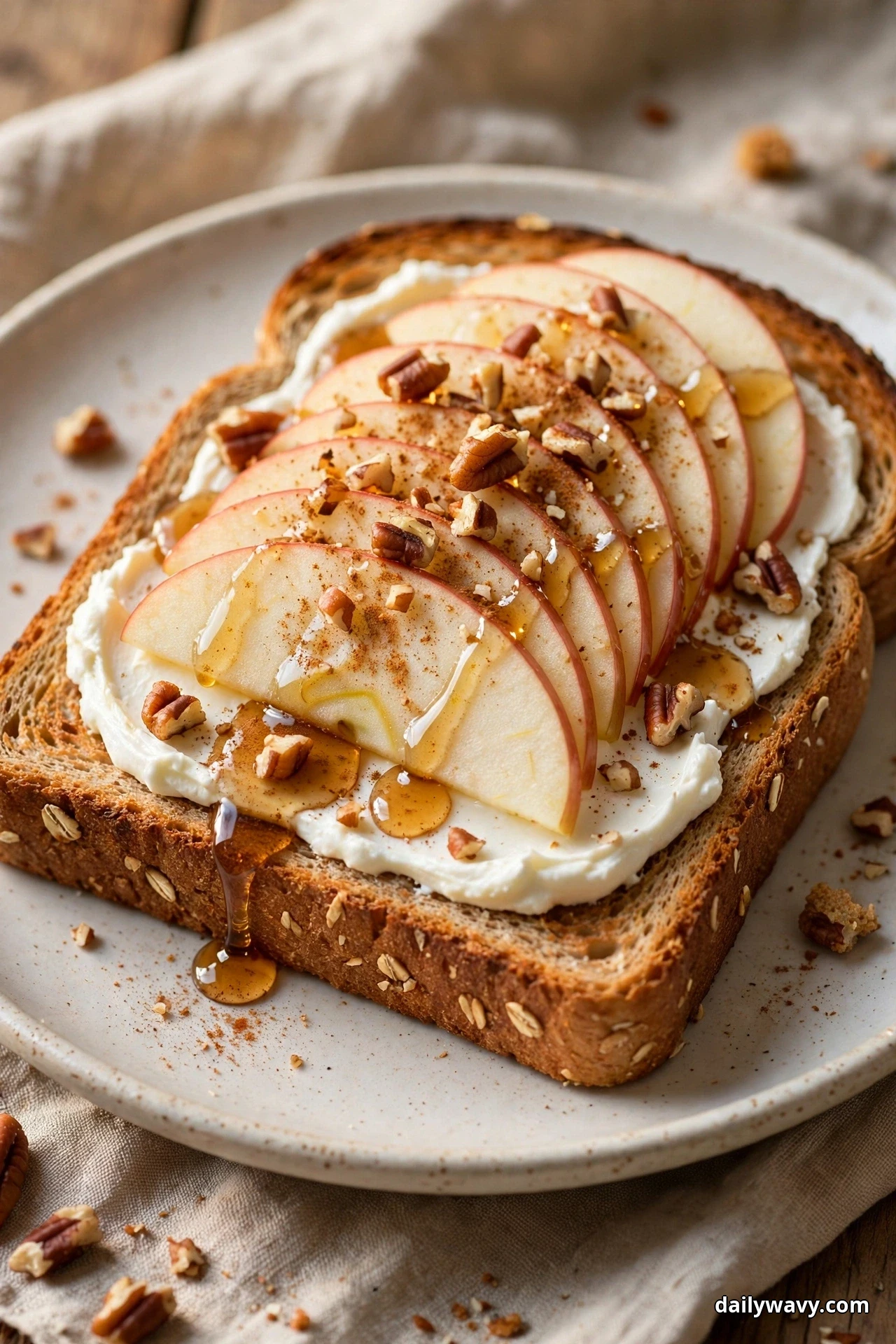 A slice of multigrain toast spread with cream cheese is topped with fanned apple slices, a maple syrup drizzle, a dusting of cinnamon and chopped pecans