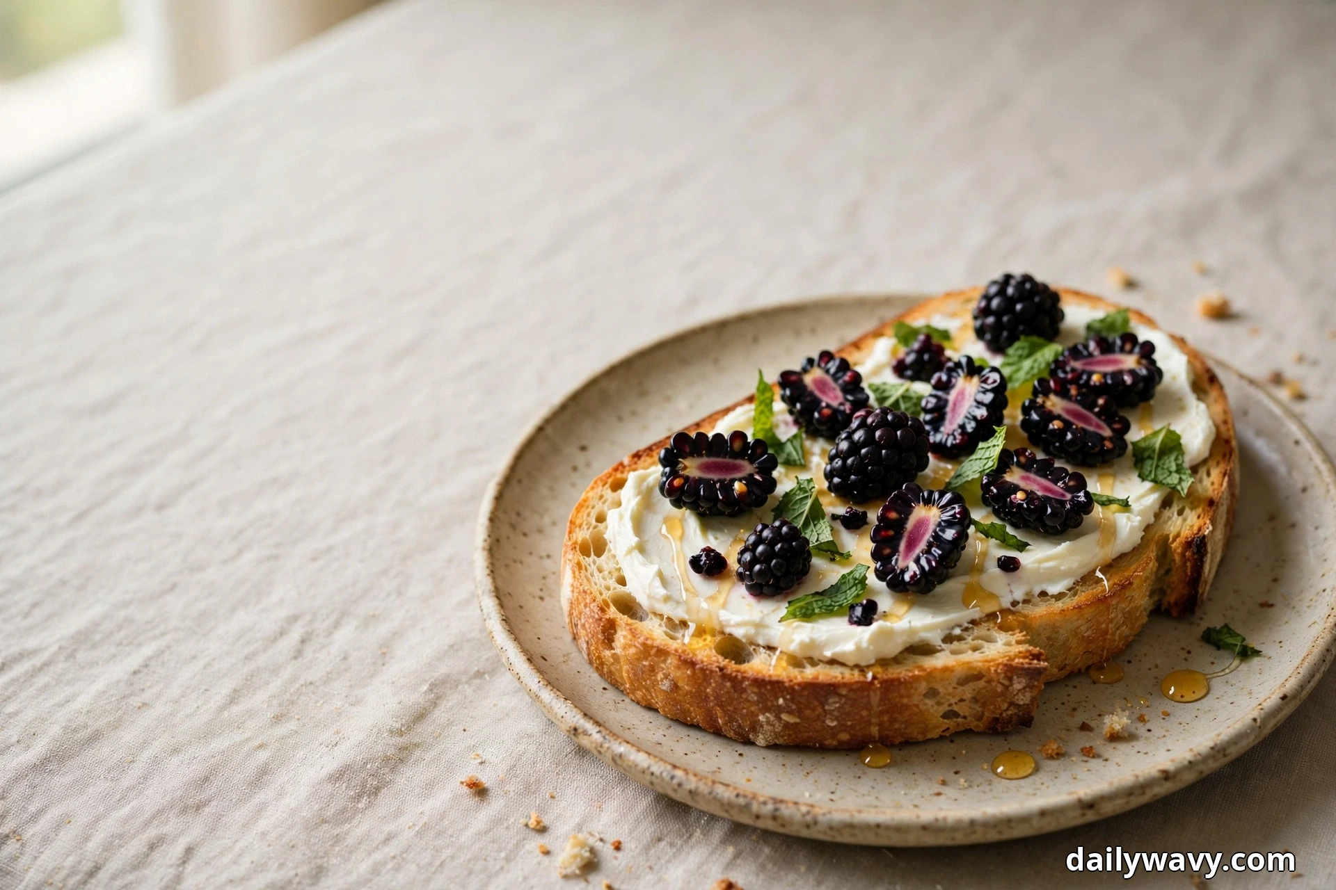 A slice of sourdough toast spread with cream cheese and topped with whole and halved blackberries, torn mint leaves and a honey drizzle rests on a speckled plate over a pale linen cloth