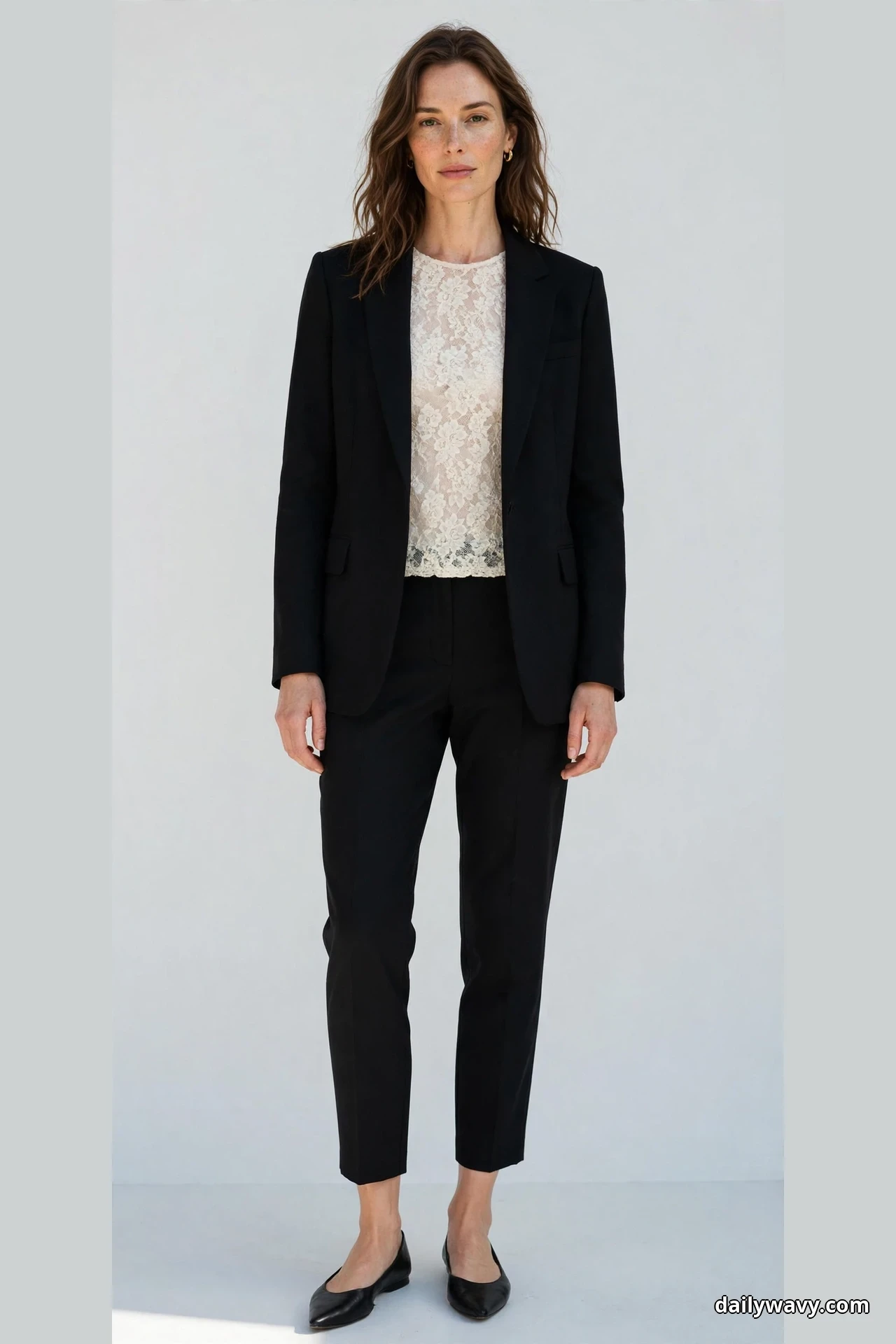A woman stands full length wearing an ivory lace top under a tailored black blazer worn open with black tapered cropped trousers and black pointed flats below the hems
