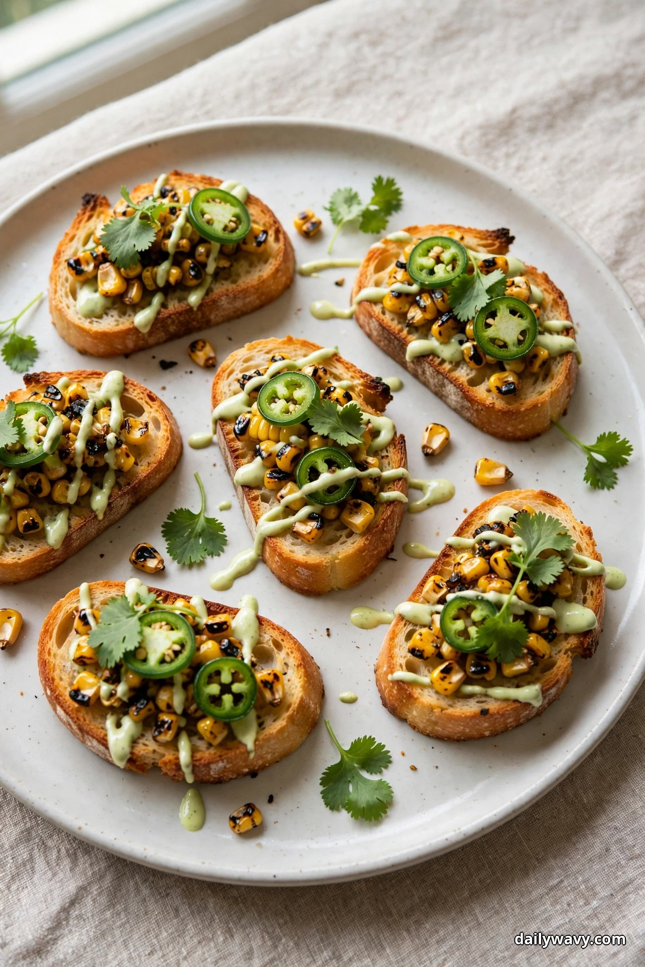 Crostini topped with charred corn kernels, jalapeño slices, a lime crema drizzle and cilantro leaves sit on a plate