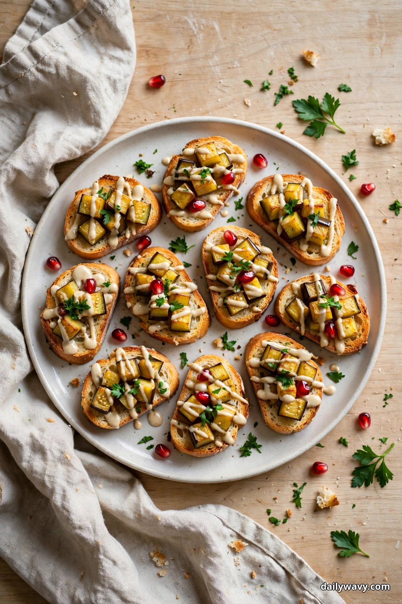 Crostini topped with roasted eggplant cubes, a tahini drizzle, pomegranate seeds and chopped parsley sit on a round plate