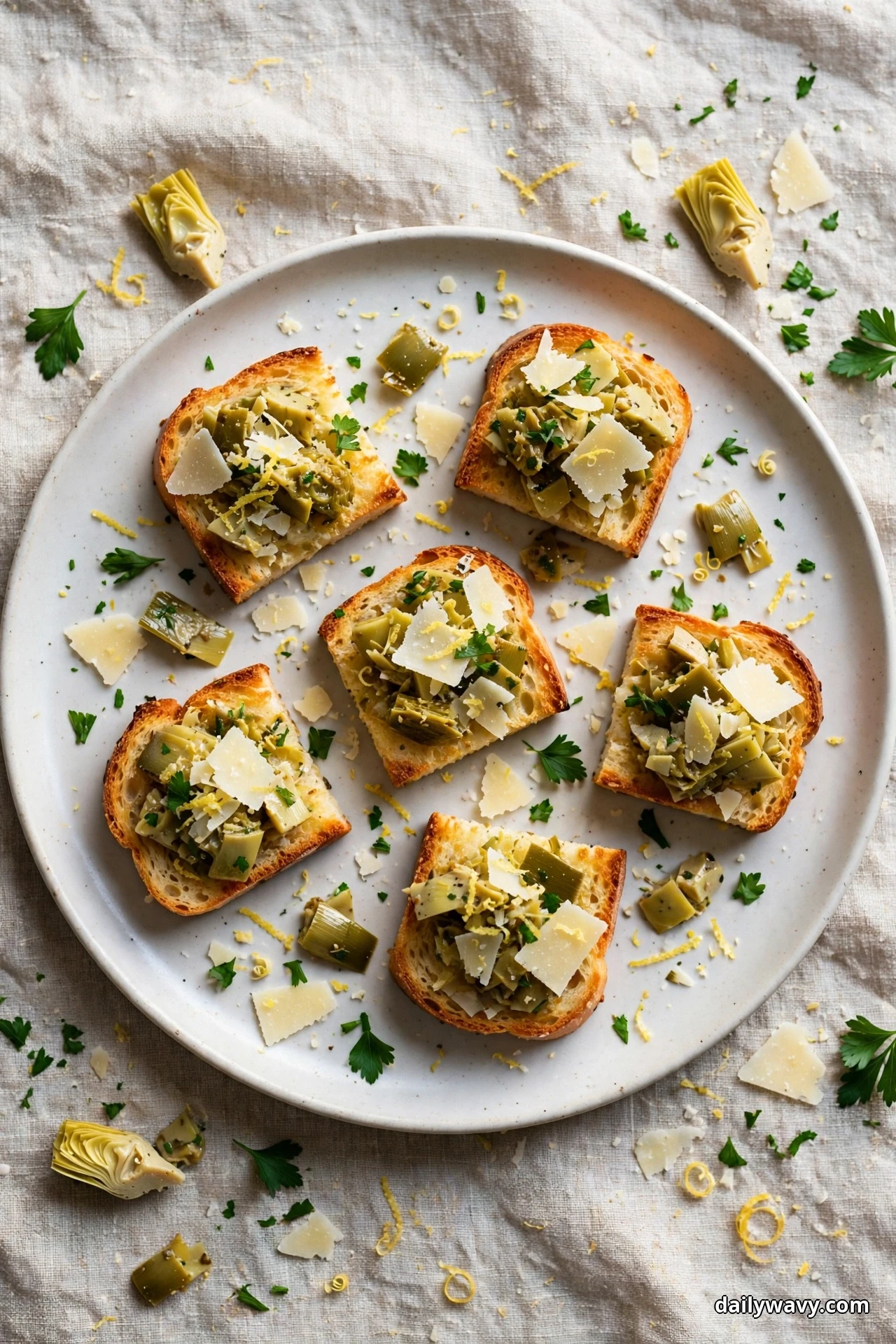 Crostini topped with chopped marinated artichoke hearts, shaved parmesan, lemon zest and parsley sit on a plate