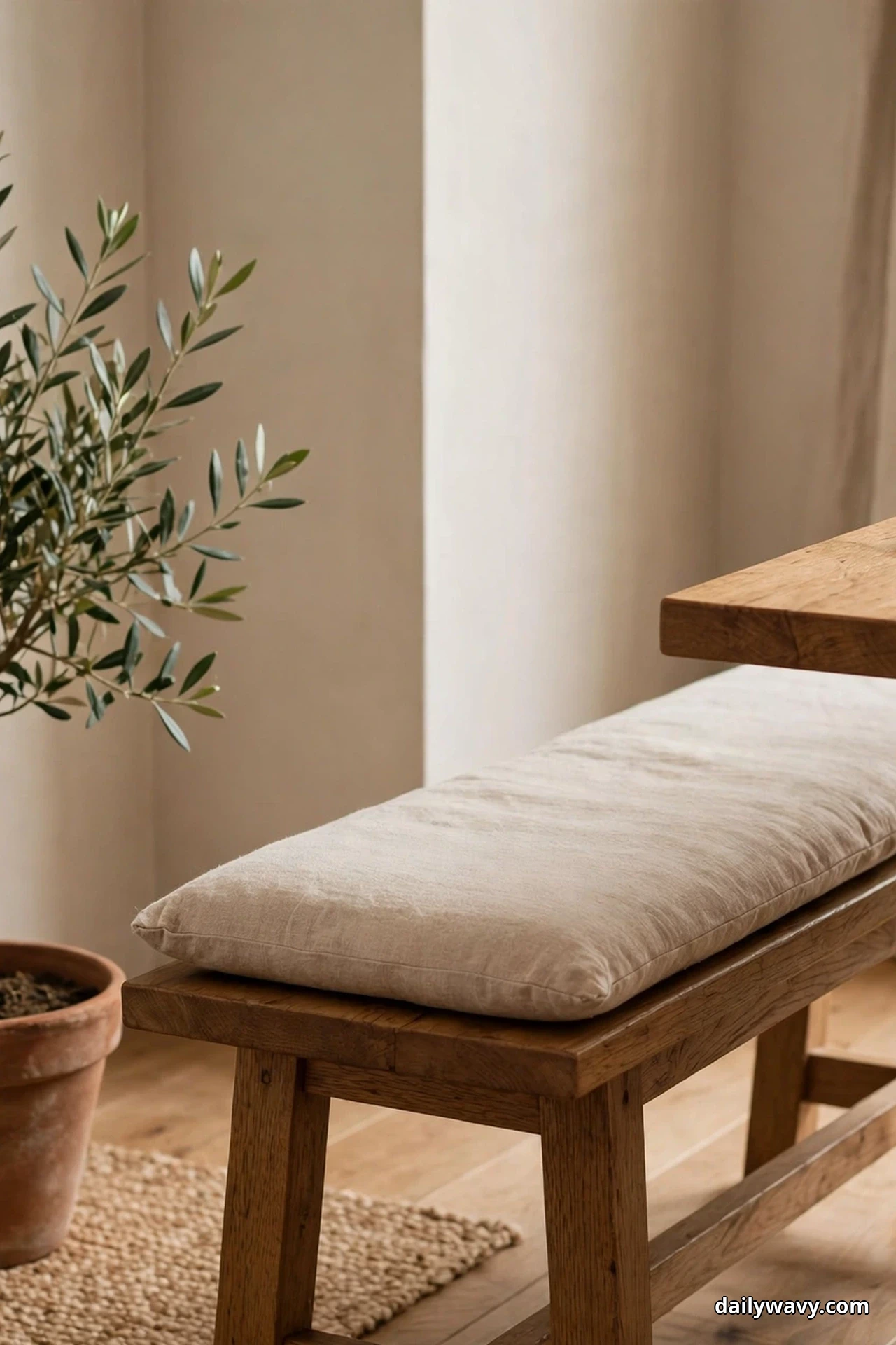 A wooden bench with a long cream linen cushion sits beside the corner of an oak table, with a potted olive tree in a terracotta pot at its end