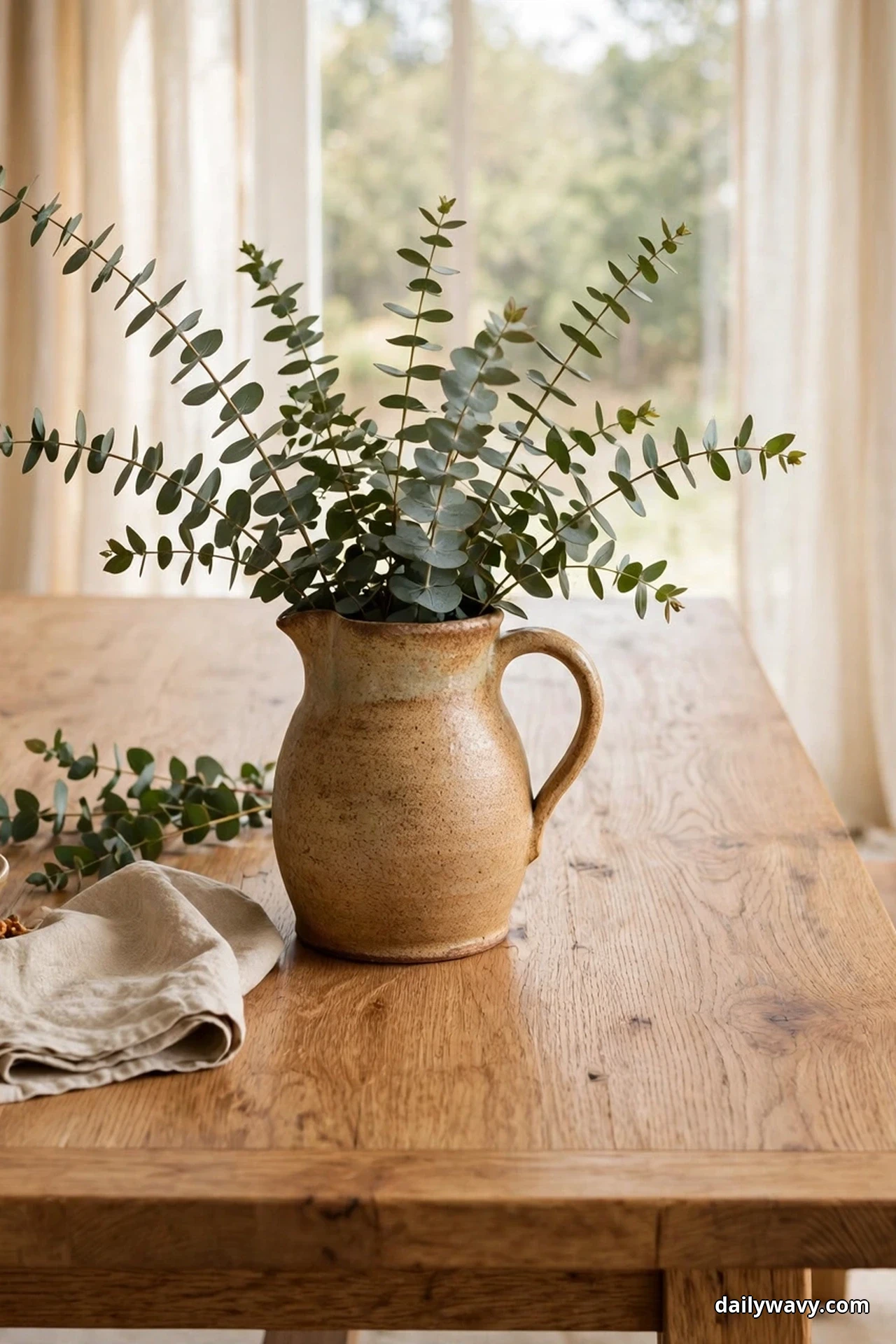 A handmade ceramic pitcher filled with eucalyptus stems stands on an oak dining table beside a folded linen cloth and a loose eucalyptus sprig