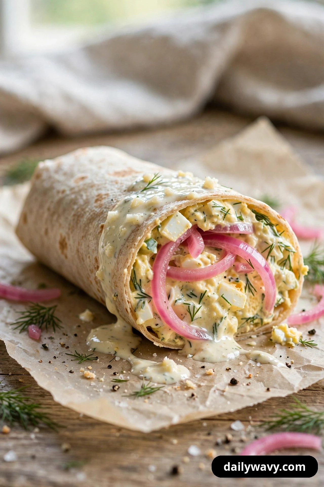 An egg salad wrap half on parchment is cut open to show pink pickled onion rings in the filling, with creamy sauce drizzled over the top and dill scattered around it.
