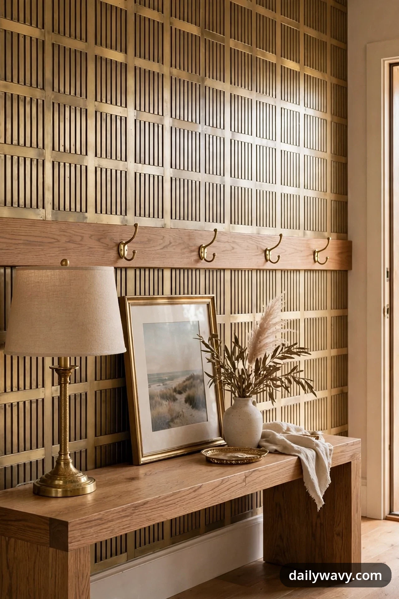 Brass repeats across an entryway scene in the hooks on a wooden wall rail, a table lamp, a leaning picture frame, and a small tray on a wooden bench, against a brass-toned lattice wall.