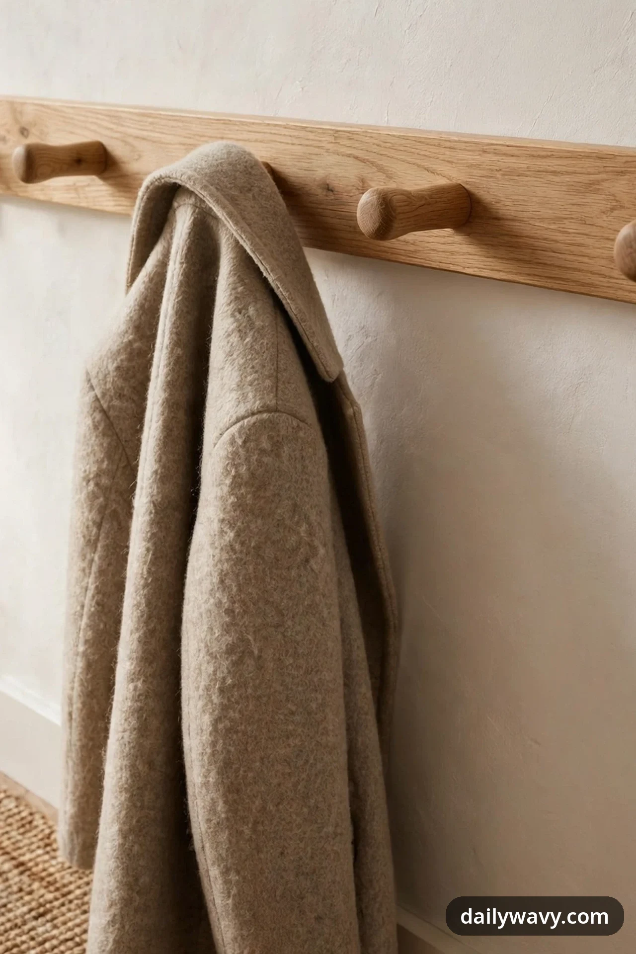 A beige wool coat hangs from one peg of a wooden peg rail, with empty pegs spaced widely on either side.