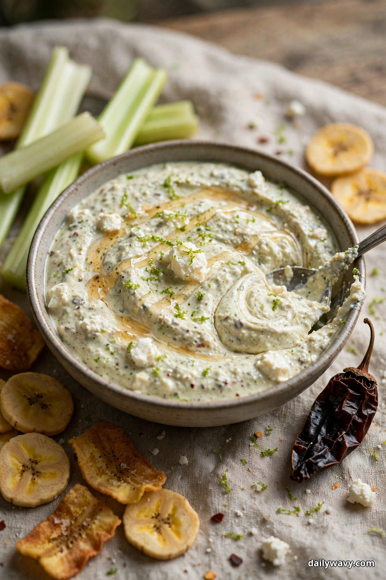 A bowl of speckled chipotle feta dip with a honey drizzle and lime zest sits with plantain chips and celery sticks