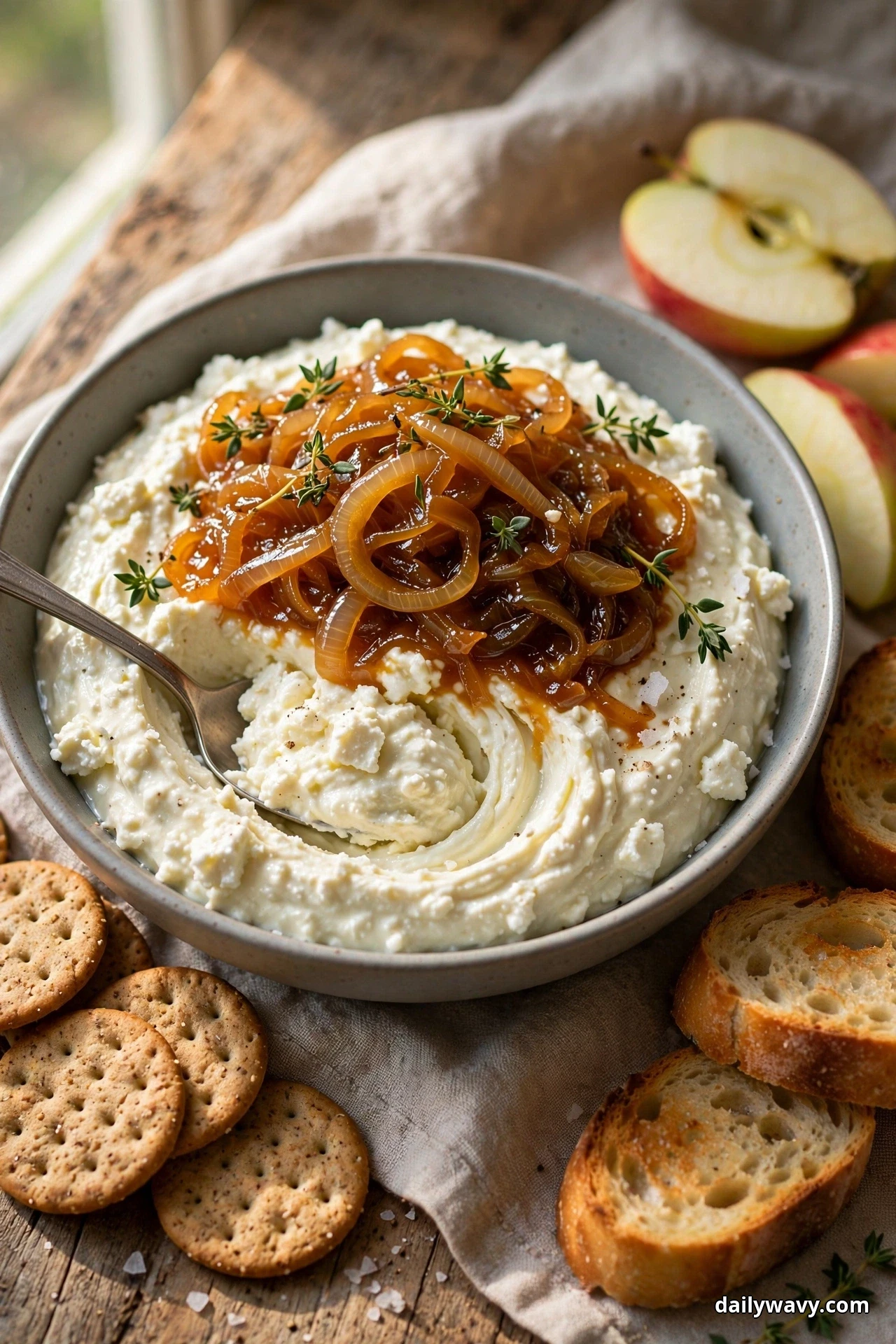 A bowl of feta dip piled with jammy caramelized onions and thyme sprigs sits with sturdy crackers, toasted baguette and apple slices