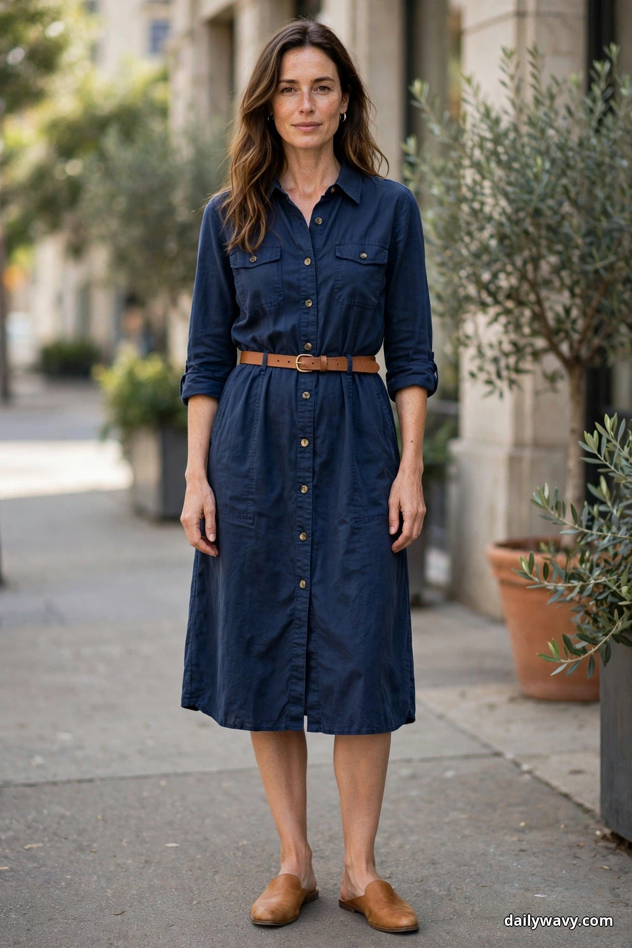 A woman stands full length on a city sidewalk wearing a navy shirt dress with a narrow tan leather belt at the waist and sleeves rolled to the forearm, with tan leather flat mules
