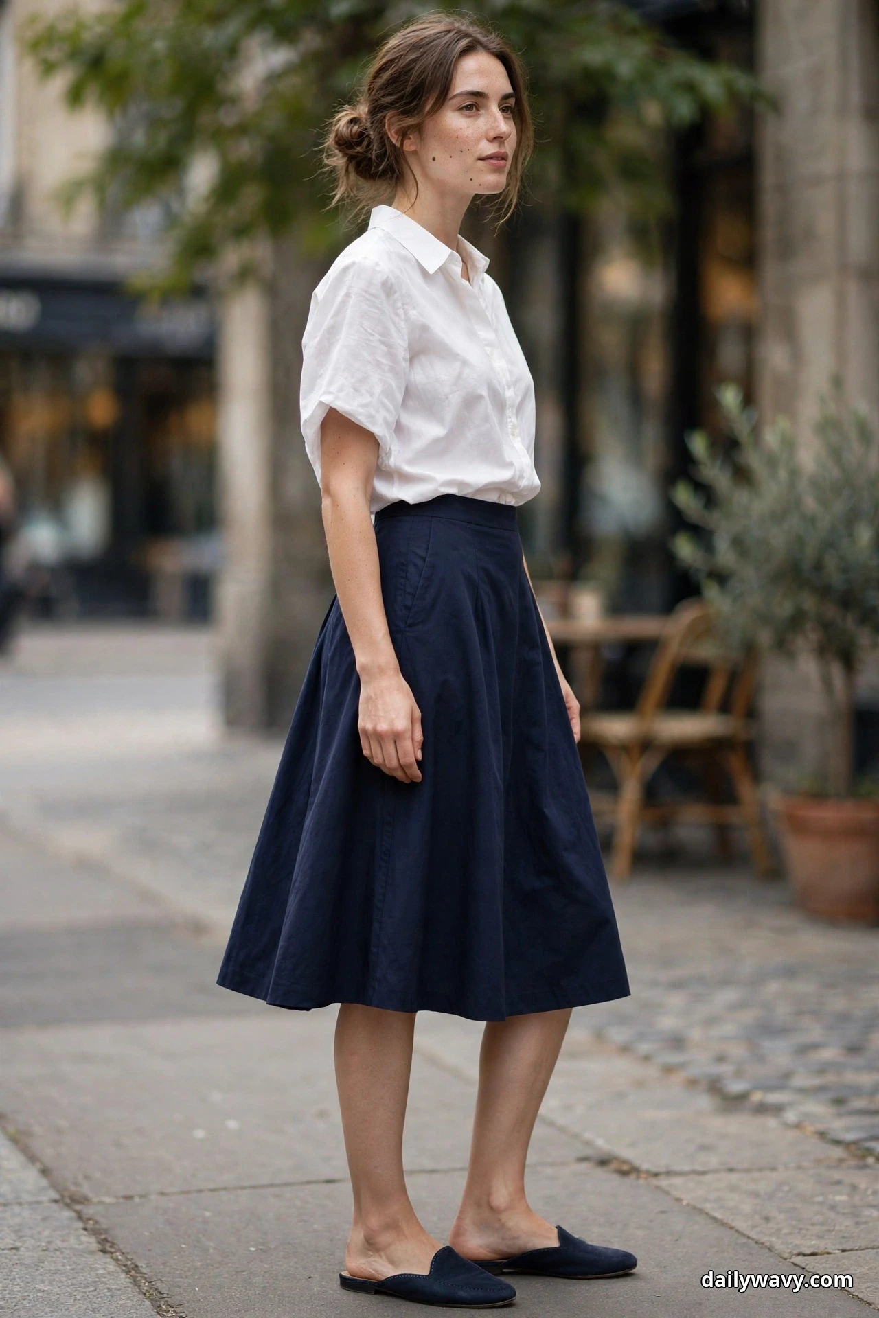 A woman stands in a three-quarter pose on a cafe street wearing a white cotton blouse tucked into a navy A-line skirt that falls below the knee, with navy suede flat mules