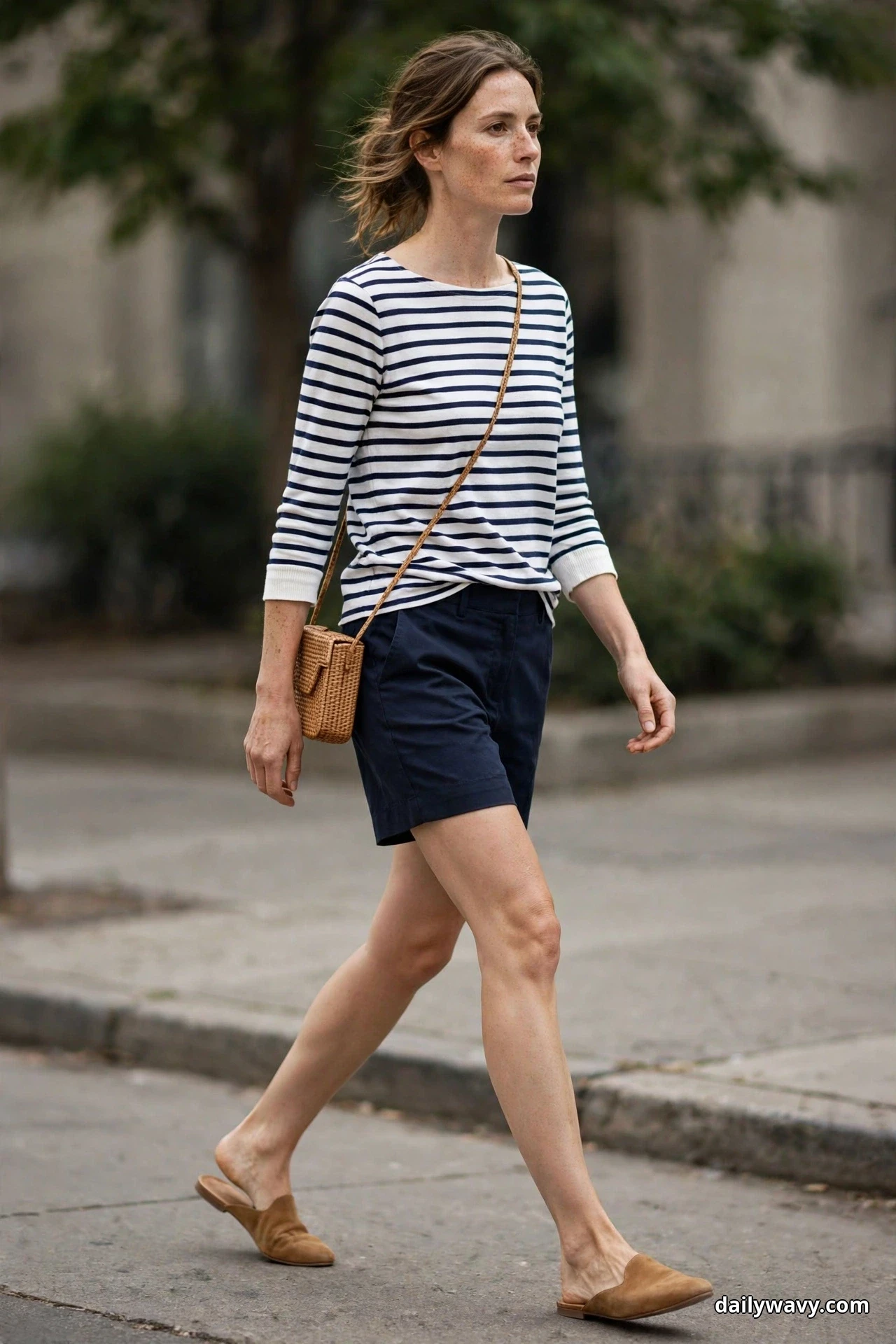 A woman walks along a city street wearing a navy and white striped Breton top half-tucked into navy tailored shorts, carrying a woven crossbody bag, with tan suede flat mules