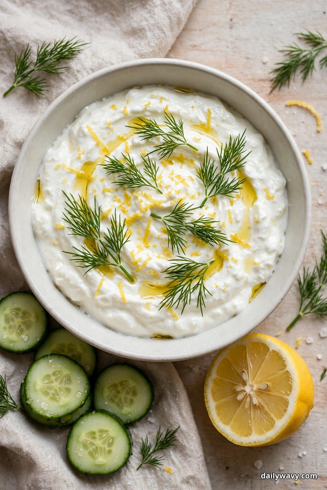 An overhead view shows a bowl of Greek yogurt dip topped with lemon zest, fresh dill fronds and an olive oil drizzle, with cucumber rounds and a lemon half beside it