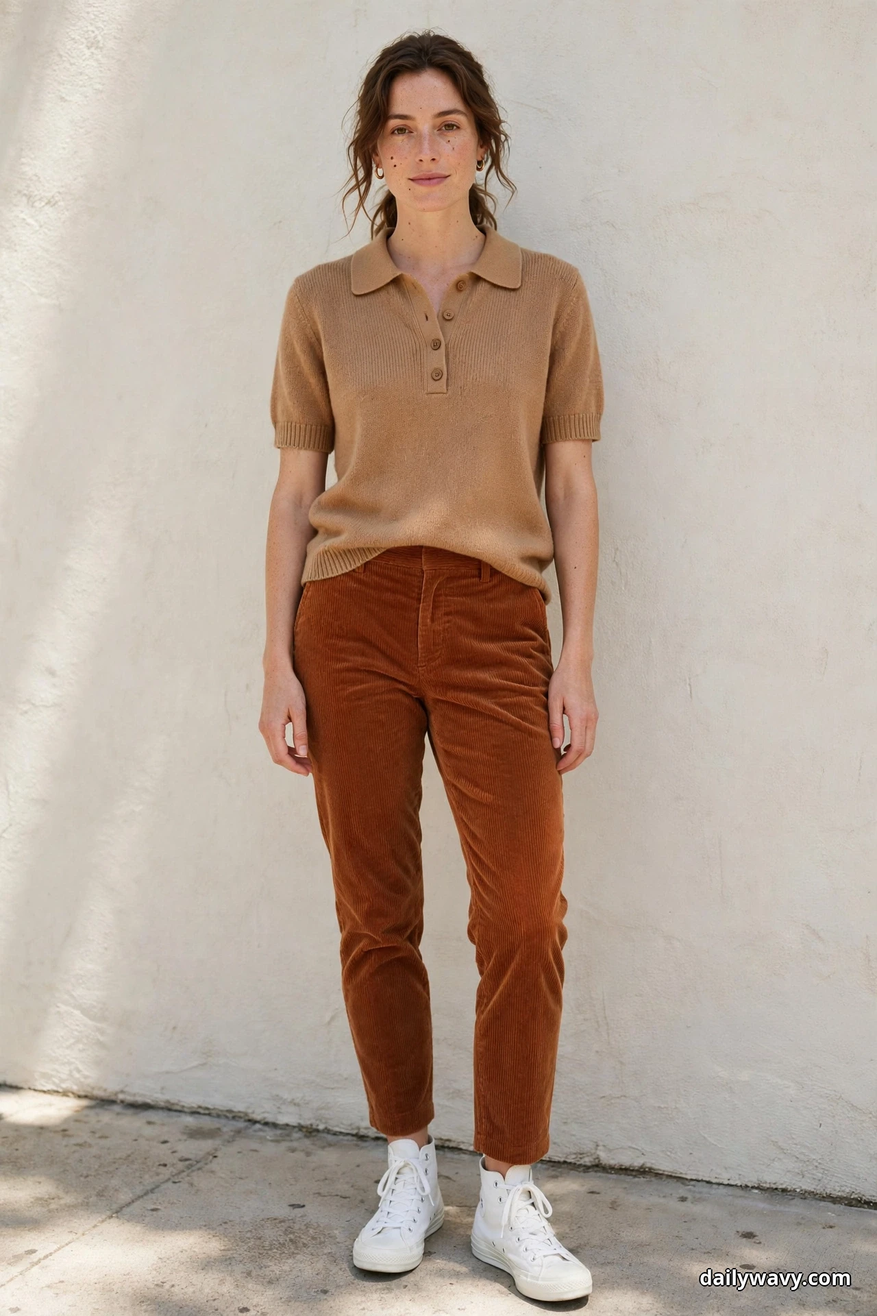 A woman wearing rust corduroy trousers, a tan knit polo and white canvas high-top sneakers stands full length against a textured wall with both sneakers showing fully below the ankle hems