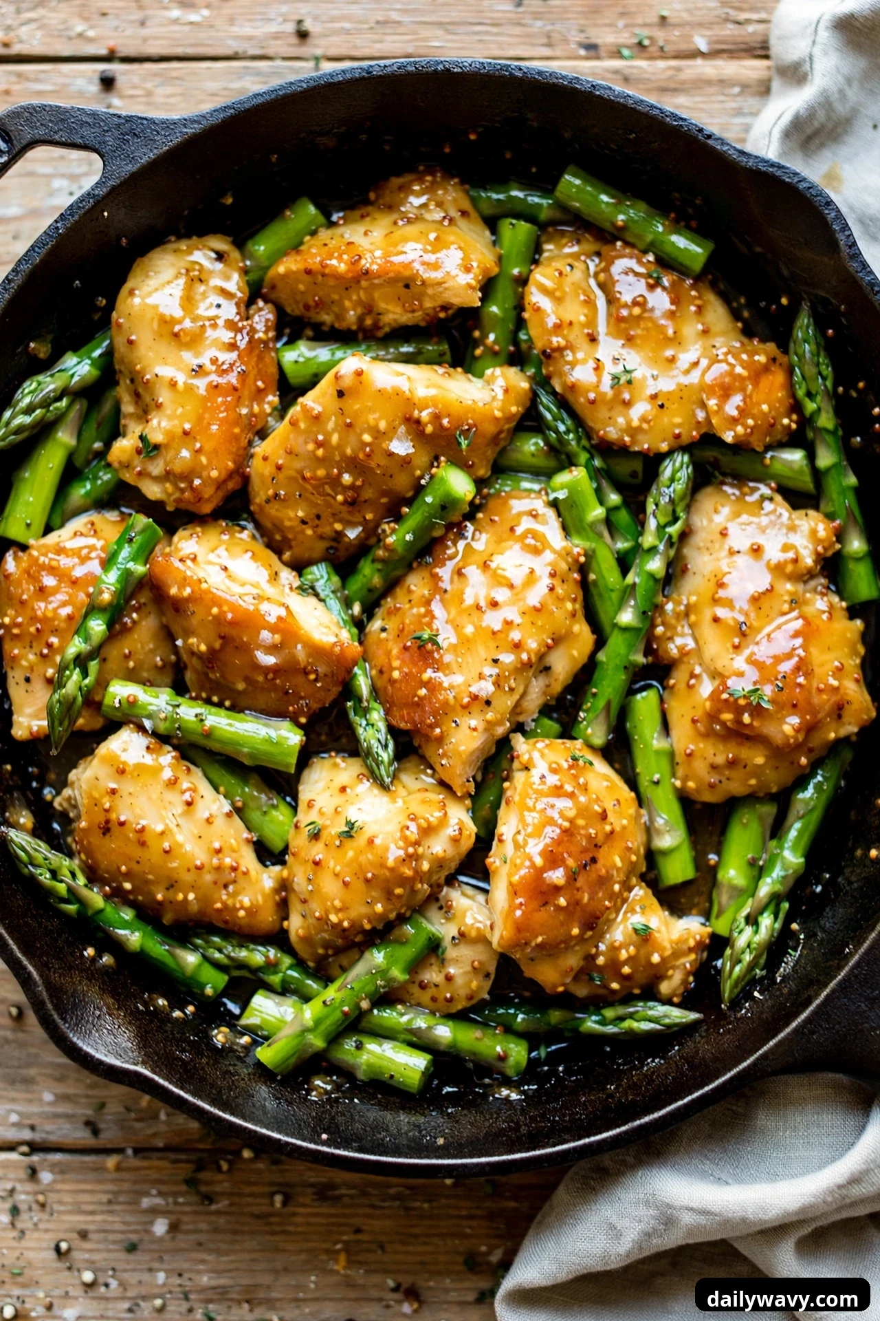 An overhead cast iron skillet holds glazed chicken pieces tucked among bright green asparagus pieces in honey mustard sauce.