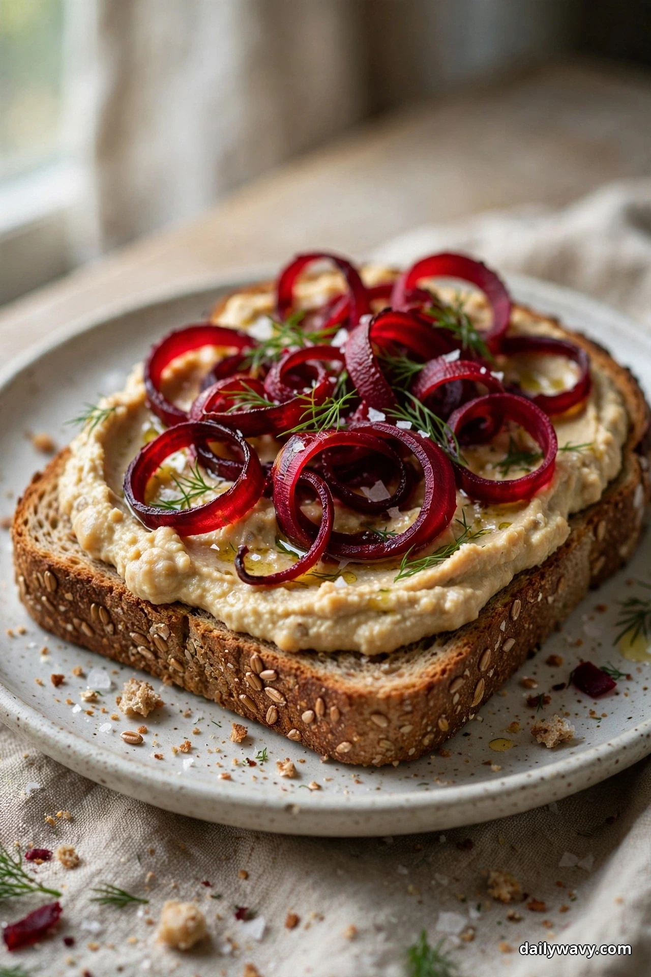 Seen up close, a thick slice of seeded toast spread with hummus is piled with curled roasted beet ribbons, dill fronds, olive oil and flaky salt on a speckled plate