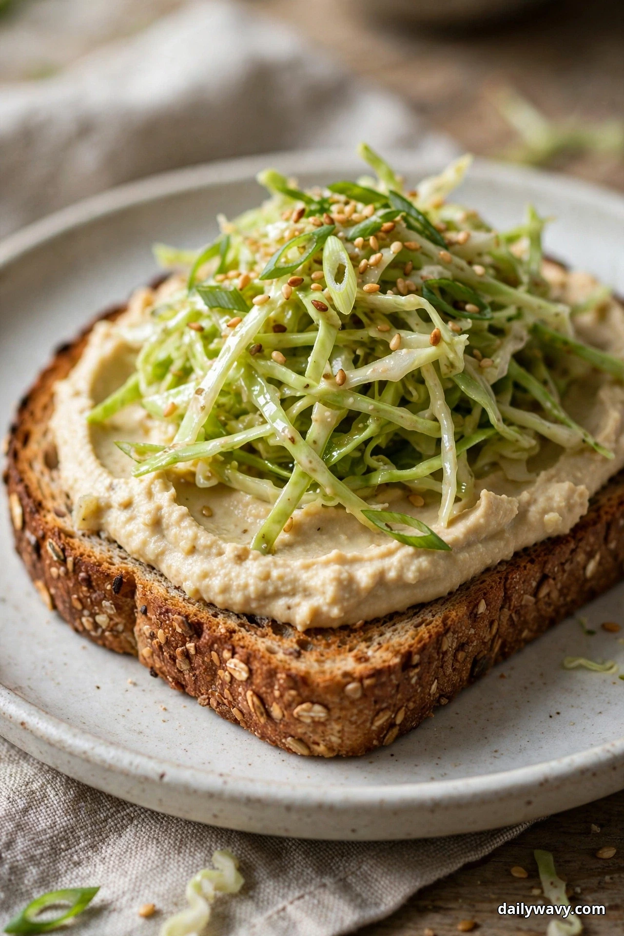 A thick slice of seeded wholegrain toast spread with hummus is mounded with shredded sesame cabbage slaw, sesame seeds and sliced scallions on a speckled plate