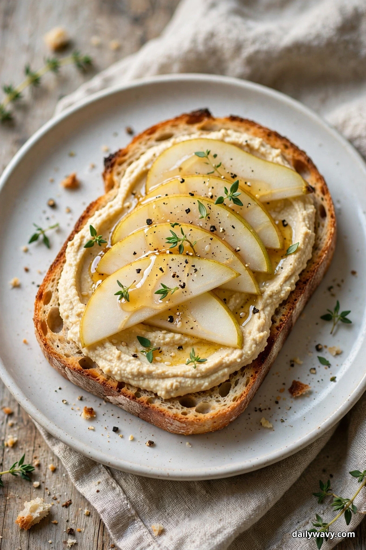 A slice of toasted sourdough spread with hummus is layered with overlapping pear slices, a drizzle of honey, thyme leaves and cracked black pepper on a white plate