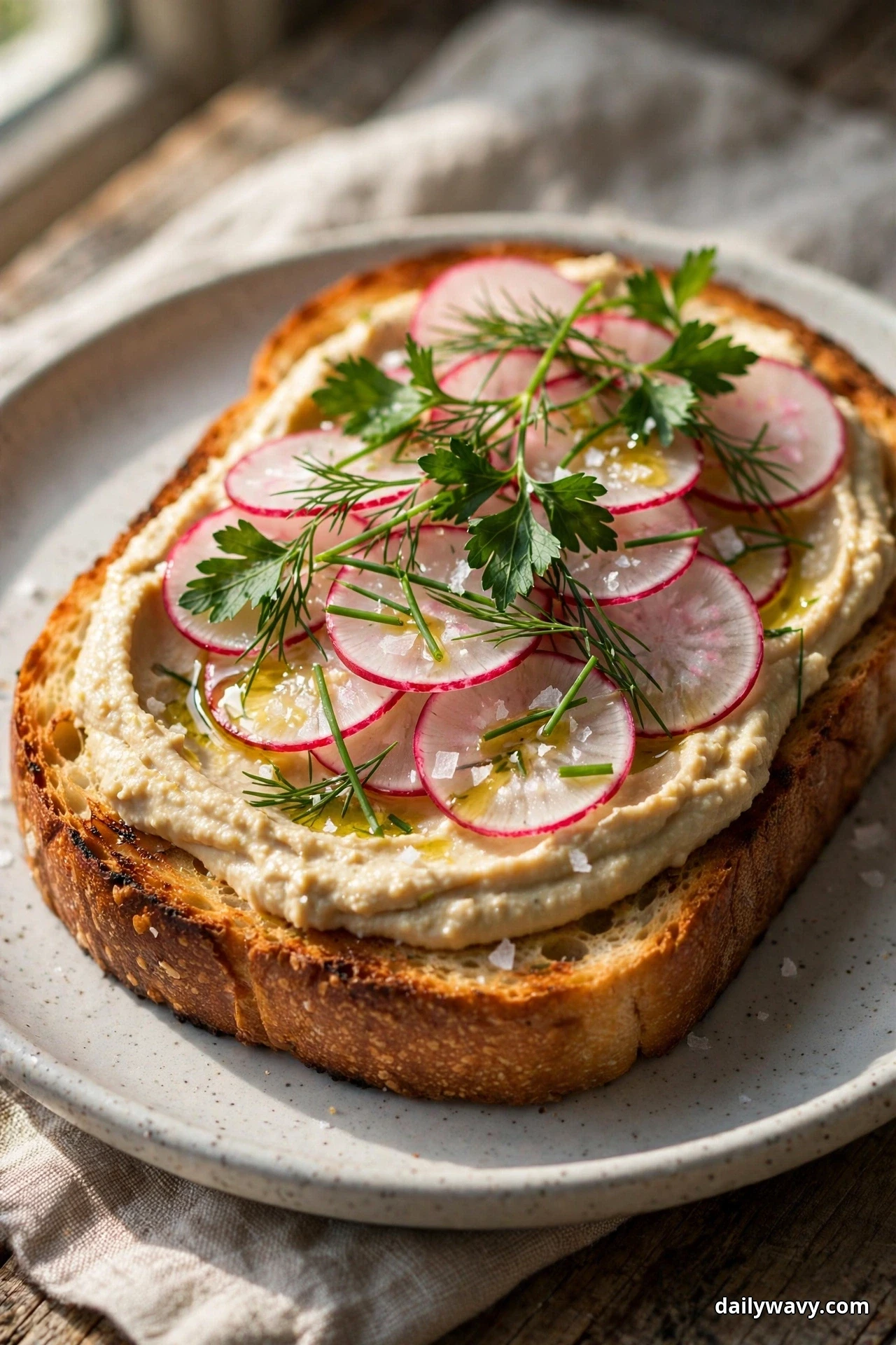 A slice of toasted bread spread with hummus is shingled with paper-thin radish slices and topped with parsley, dill, chives, olive oil and flaky salt on a speckled plate