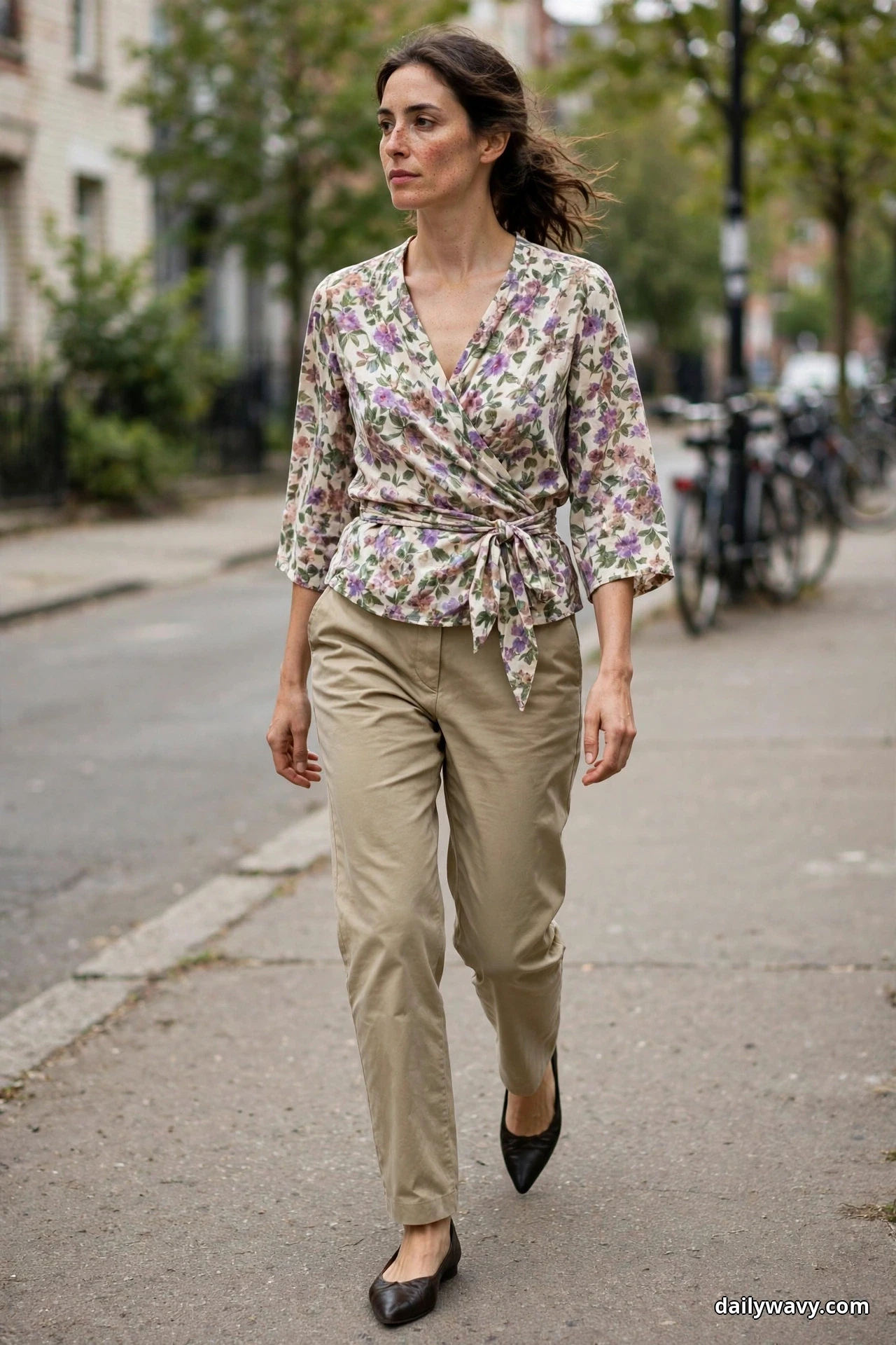 A woman walks along a sidewalk wearing a floral wrap blouse tied at the waist over slim khaki trousers, with black pointed flats below the hems