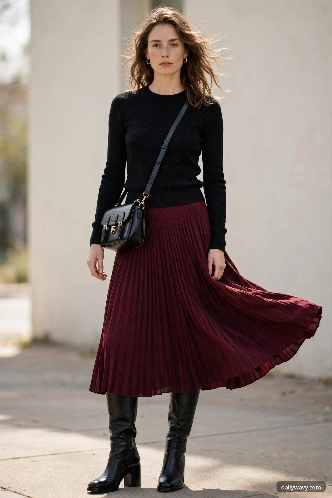 A woman in a black fine-knit sweater and burgundy pleated midi skirt with knee-high black leather boots and a black crossbody satchel, standing against a light wall.