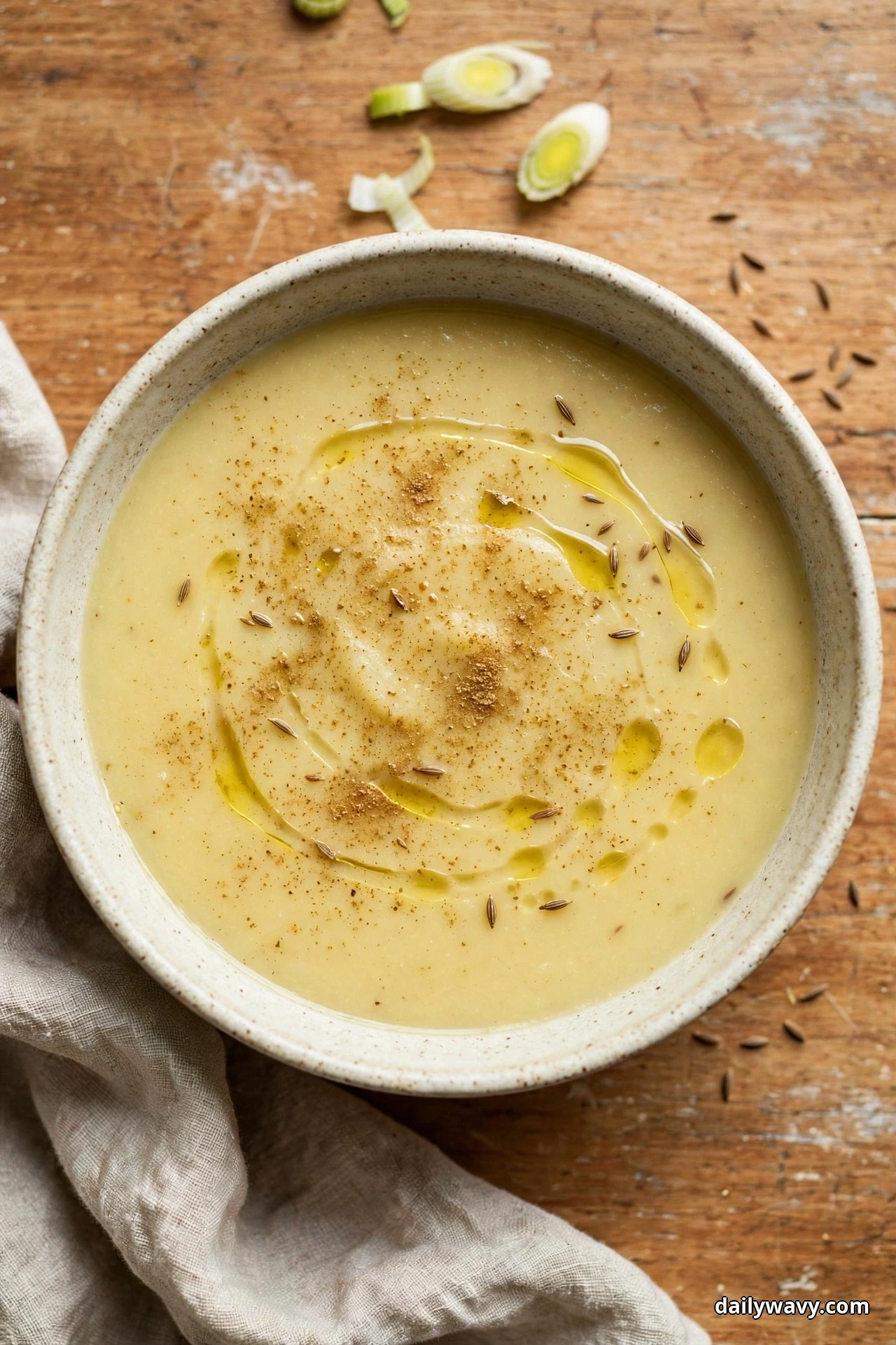 Seen from above, a speckled bowl of smooth leek and potato soup dusted with ground cumin and scattered whole cumin seeds, with swirls of olive oil on top, sits on a wooden table with sliced leek rounds beside it