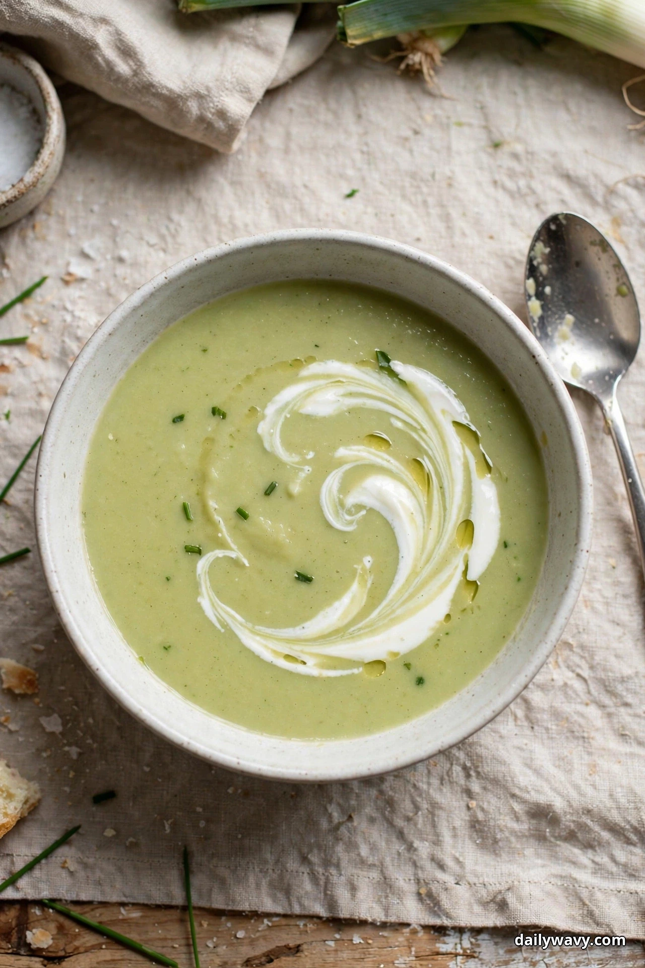 Seen from above, a speckled bowl of smooth pale green leek and potato soup with a white yogurt swirl sits on linen, a spoon resting beside the bowl