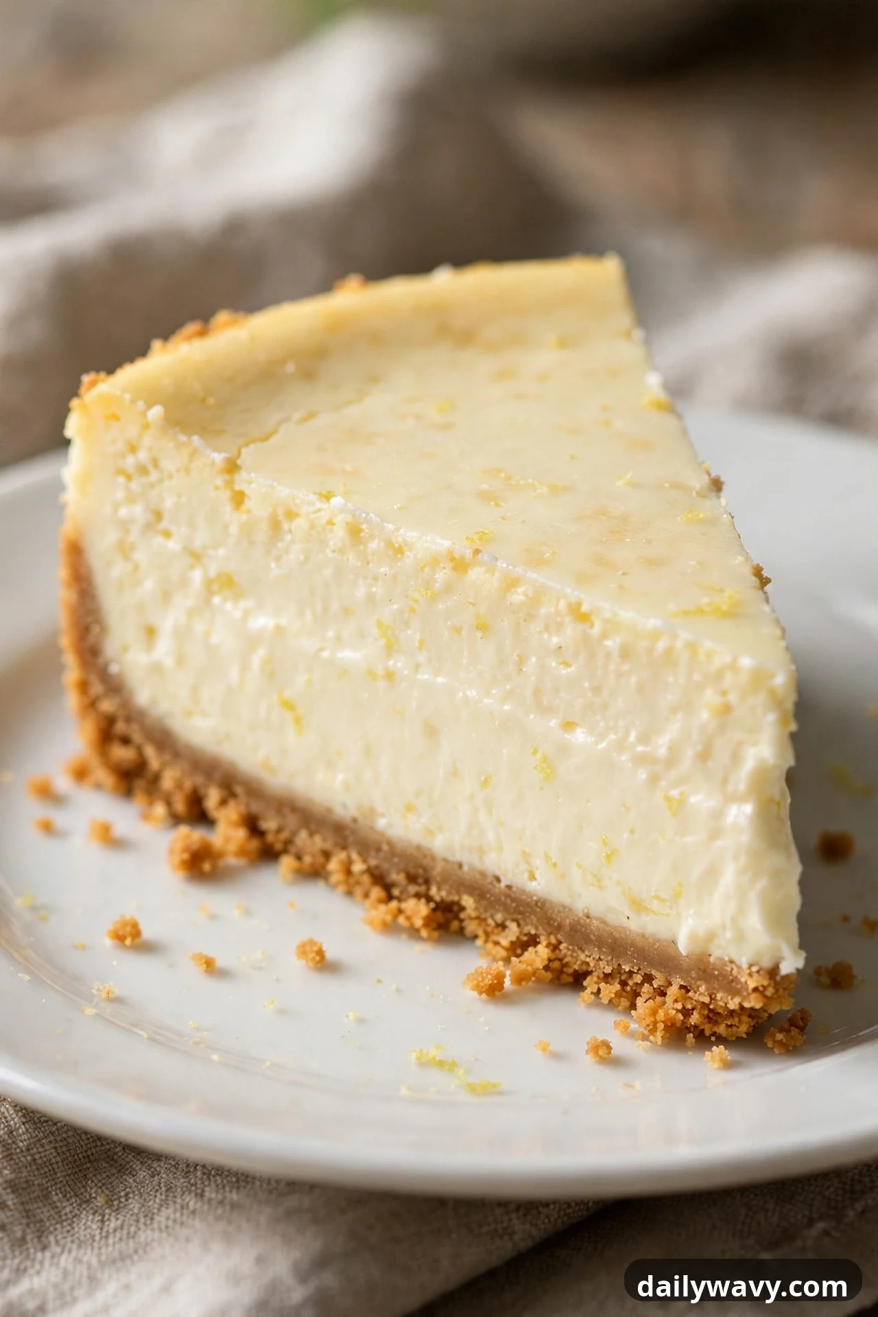 A slice of lemon mascarpone cheesecake with a smooth, glossy pale filling sits on a white plate over a golden graham crust, with fine lemon zest flecked through the filling.