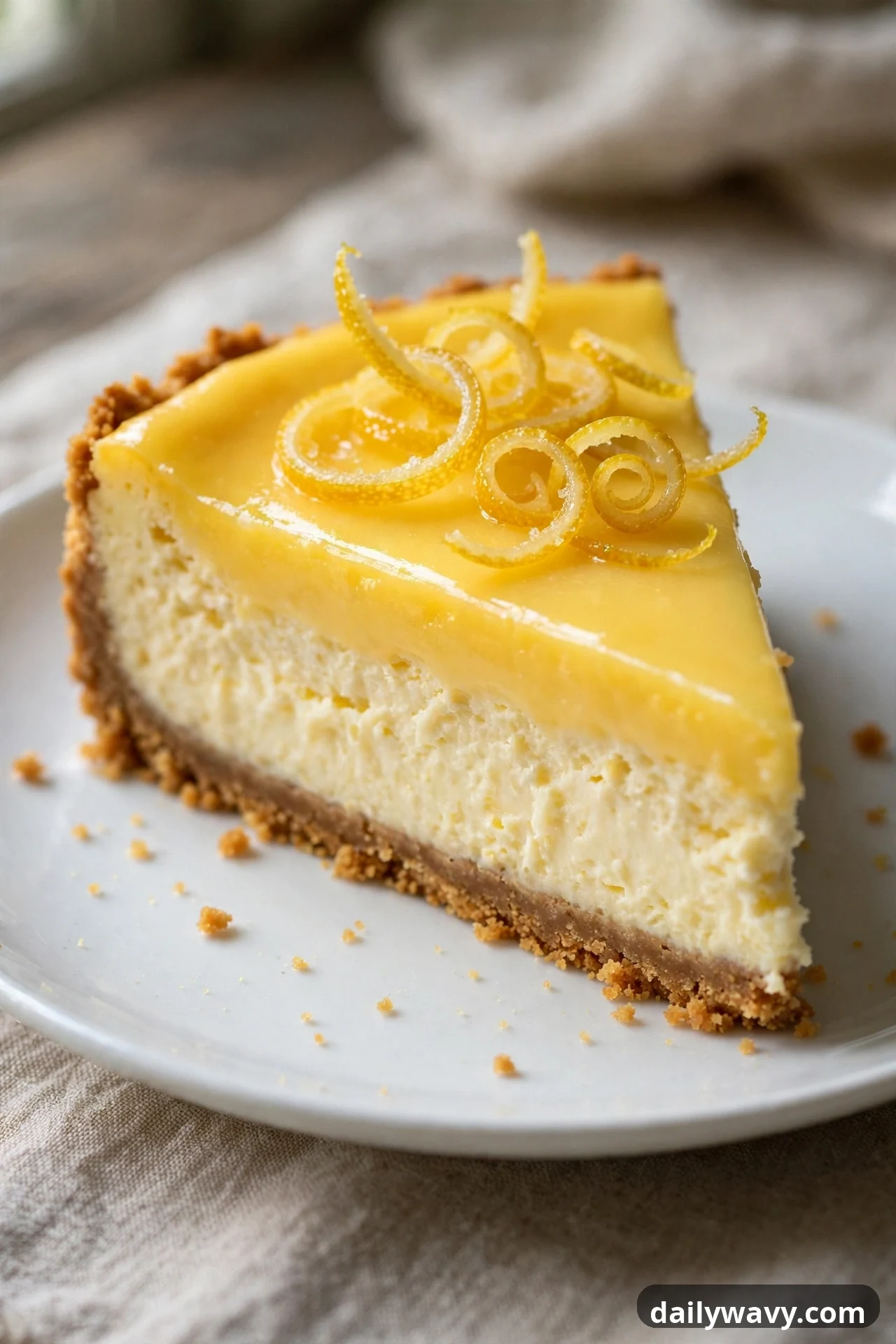 A slice of lemon cheesecake topped with a glossy layer of yellow citrus curd and candied lemon zest curls sits on a white plate over a graham crust.
