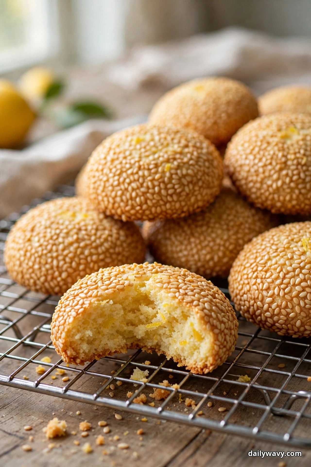 Domed lemon cookies coated all over in sesame seeds pile on a wire rack, one broken open to show a soft, fluffy center