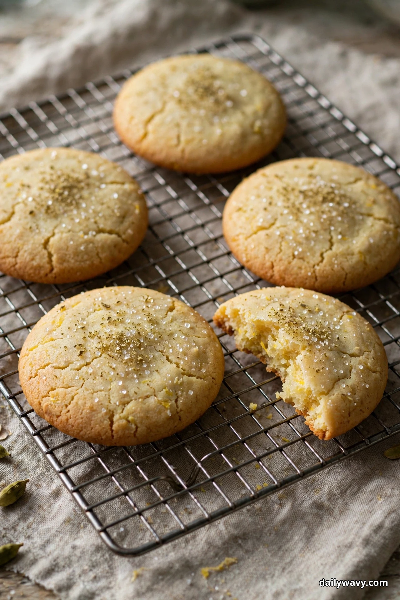 Pale lemon cookies dusted with coarse sugar and specks of cardamom cool on a wire rack, one with a bite taken out of it