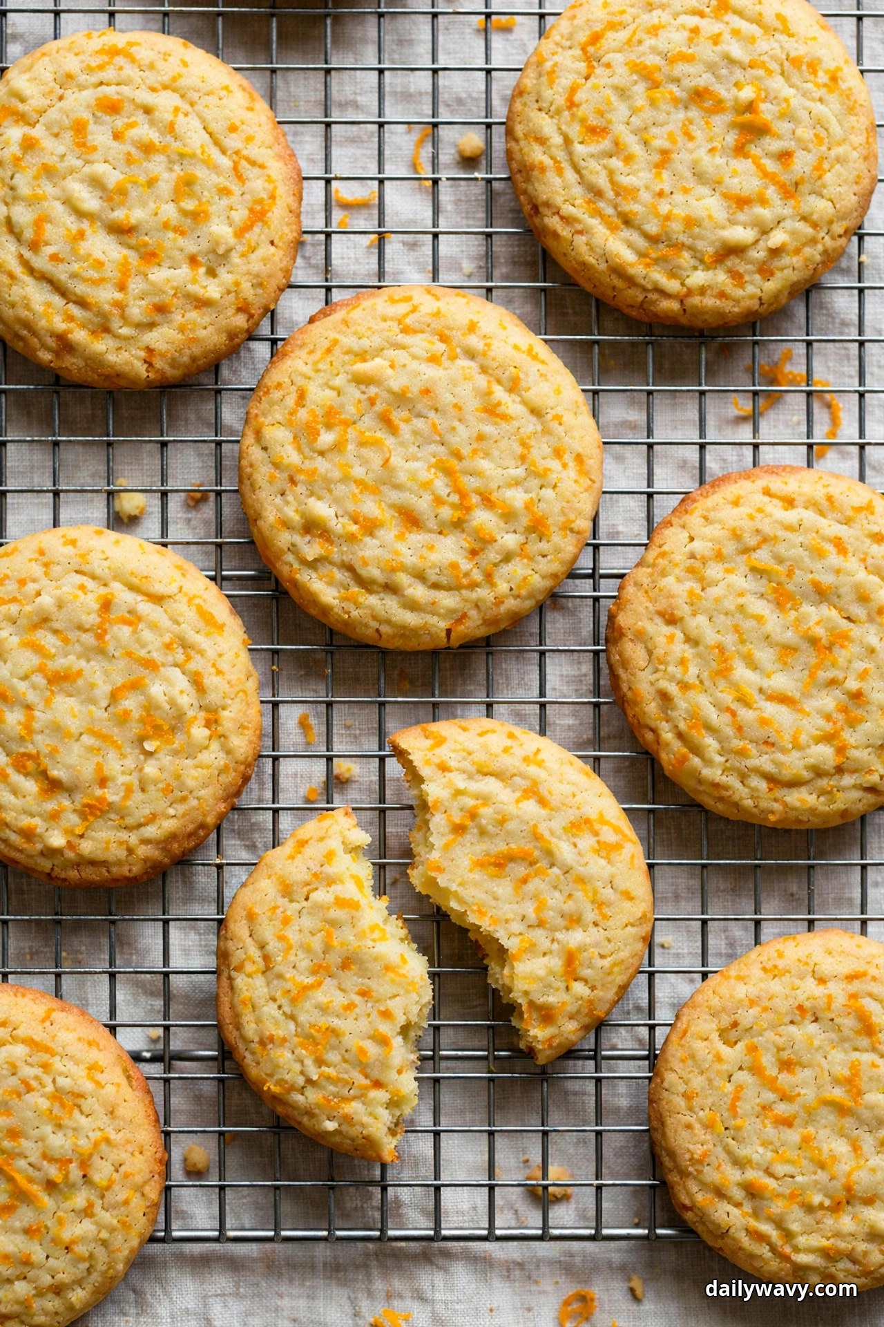 An overhead view shows lemon cookies flecked all over with orange zest cooling on a wire rack, one snapped into two halves