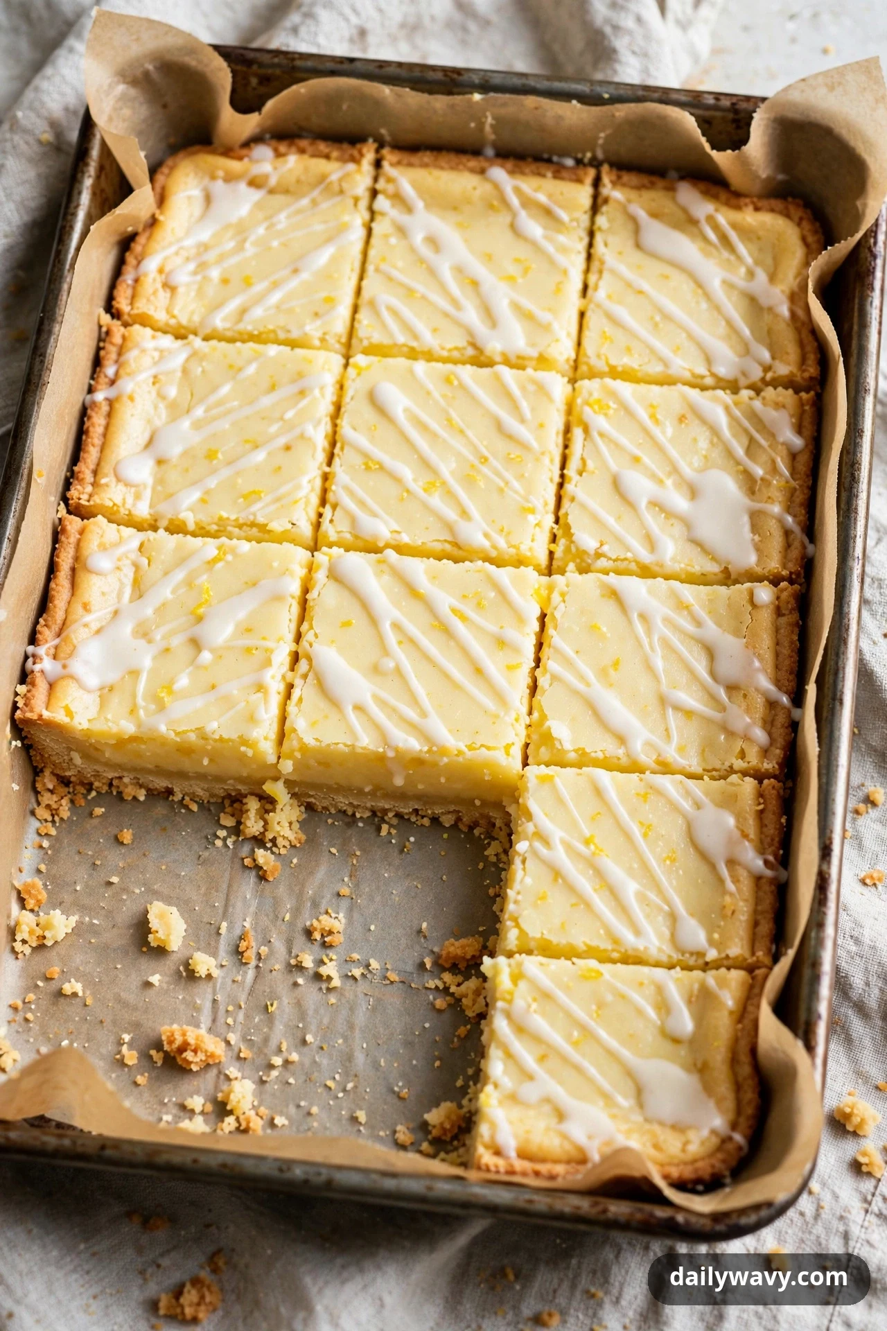 Lemon bars drizzled with white vanilla glaze in thin diagonal lines sit in a parchment-lined pan with two squares removed.