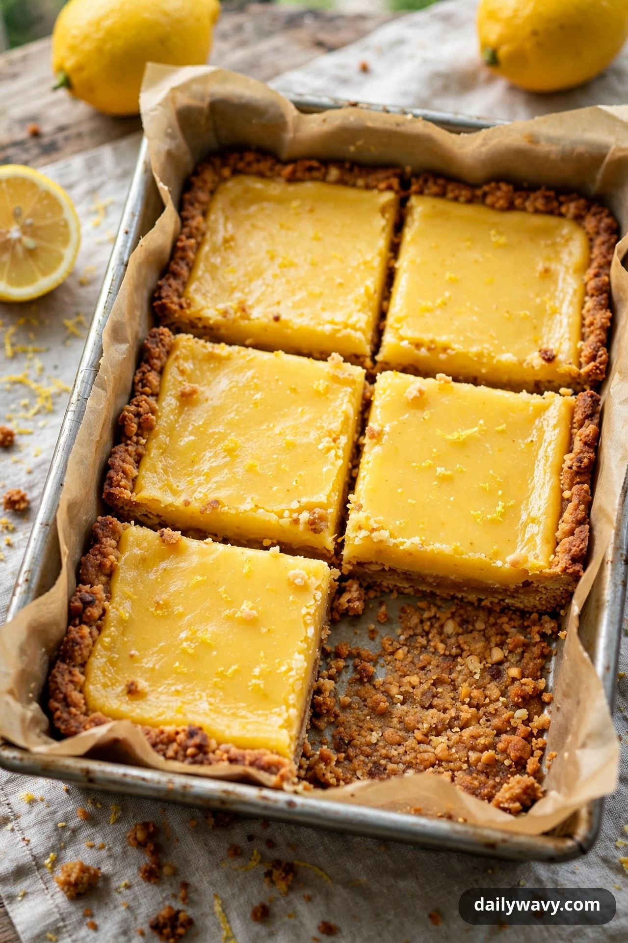 Five large lemon bar squares with deeply golden, toasty crust edges sit in a parchment-lined pan with one square removed, and whole and halved lemons rest beside the pan.
