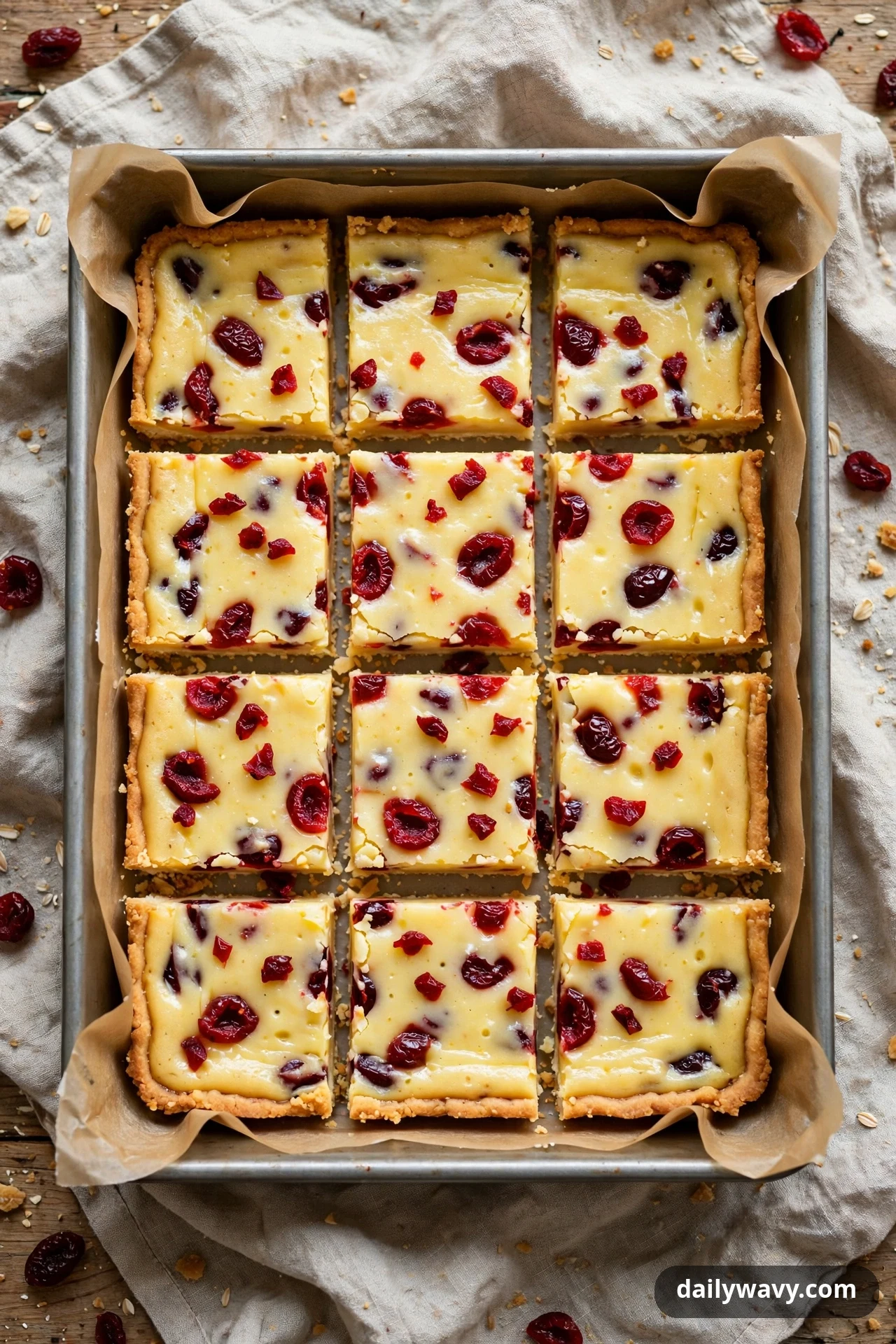 An overhead view shows twelve lemon bar squares studded with dried cherries in a parchment-lined pan, with loose dried cherries scattered on the cloth around it.