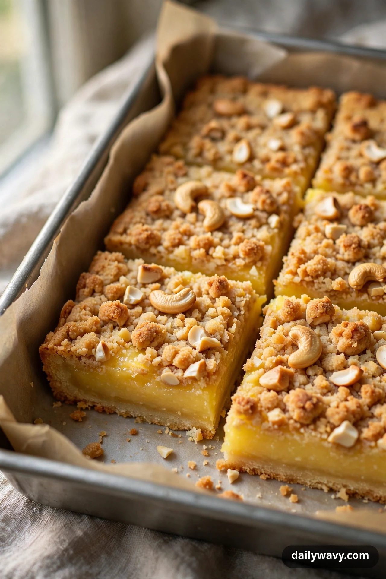 Lemon bars topped with golden cashew crumble and whole cashew pieces sit in a parchment-lined pan, with two squares pulled forward to show the layers.