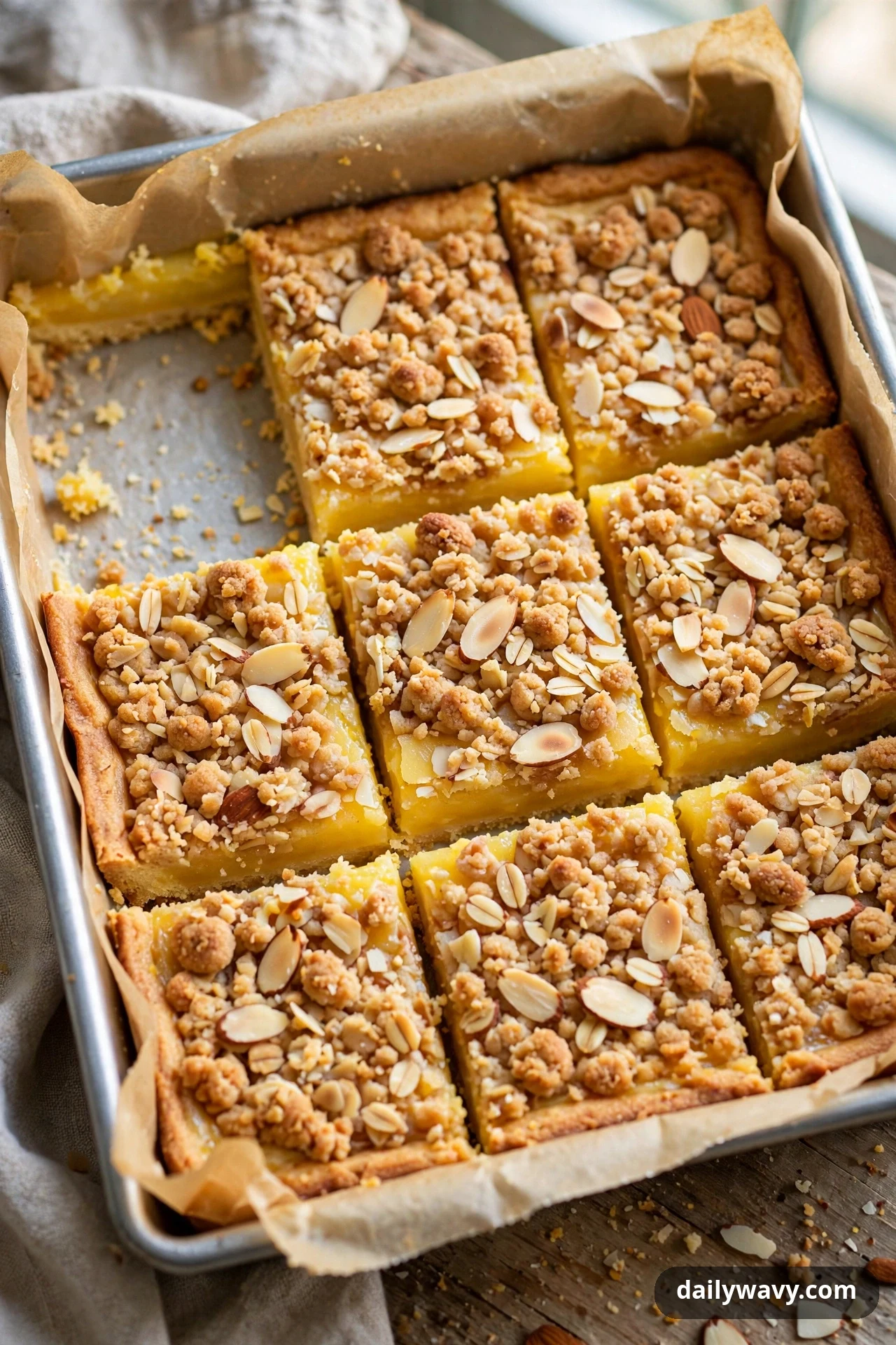 Lemon bars topped with crumble and sliced almonds sit in a parchment-lined pan with one square removed from the corner.