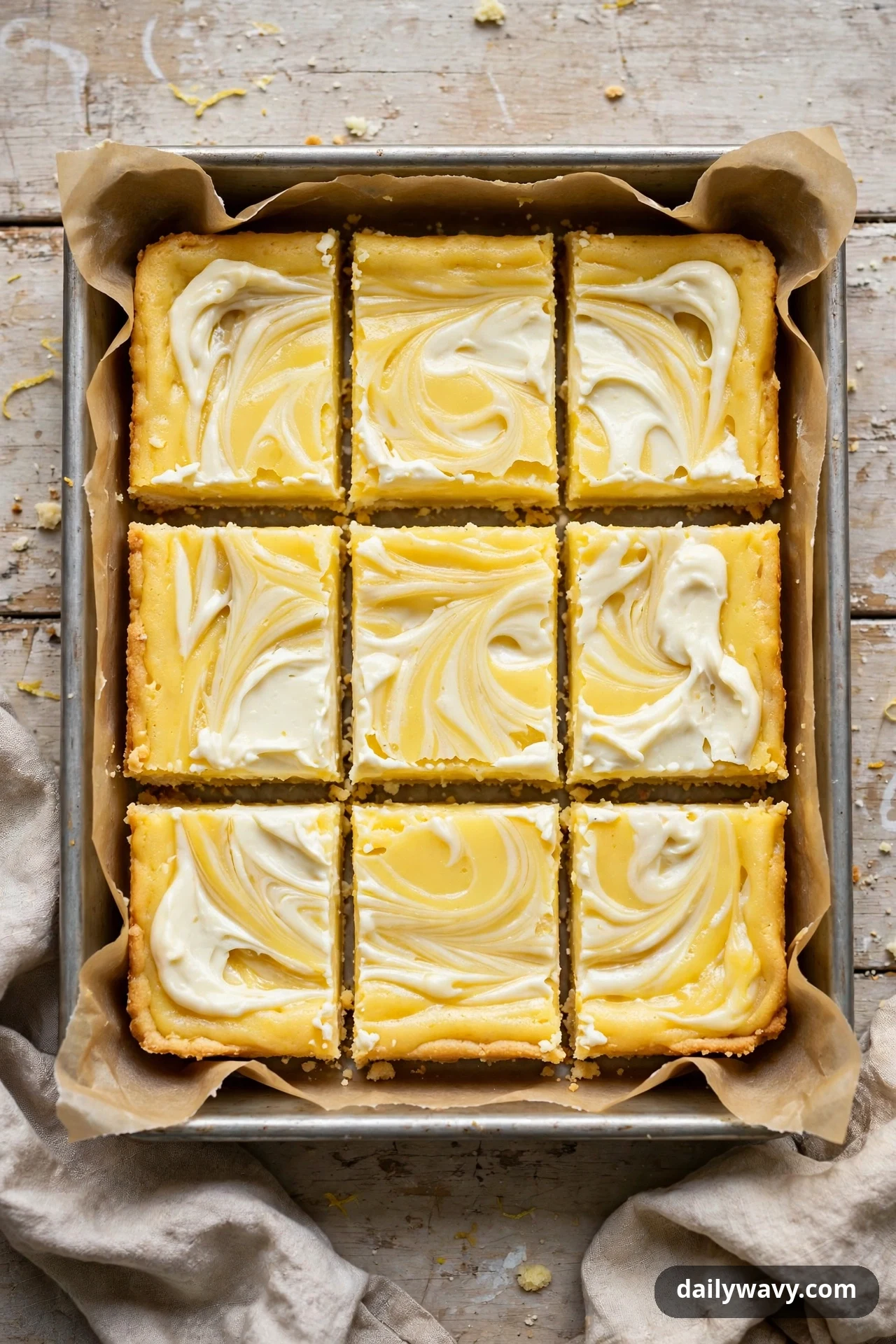 An overhead view shows nine lemon bar squares with a white cream-cheese swirl marbled through the yellow lemon layer in a parchment-lined pan.