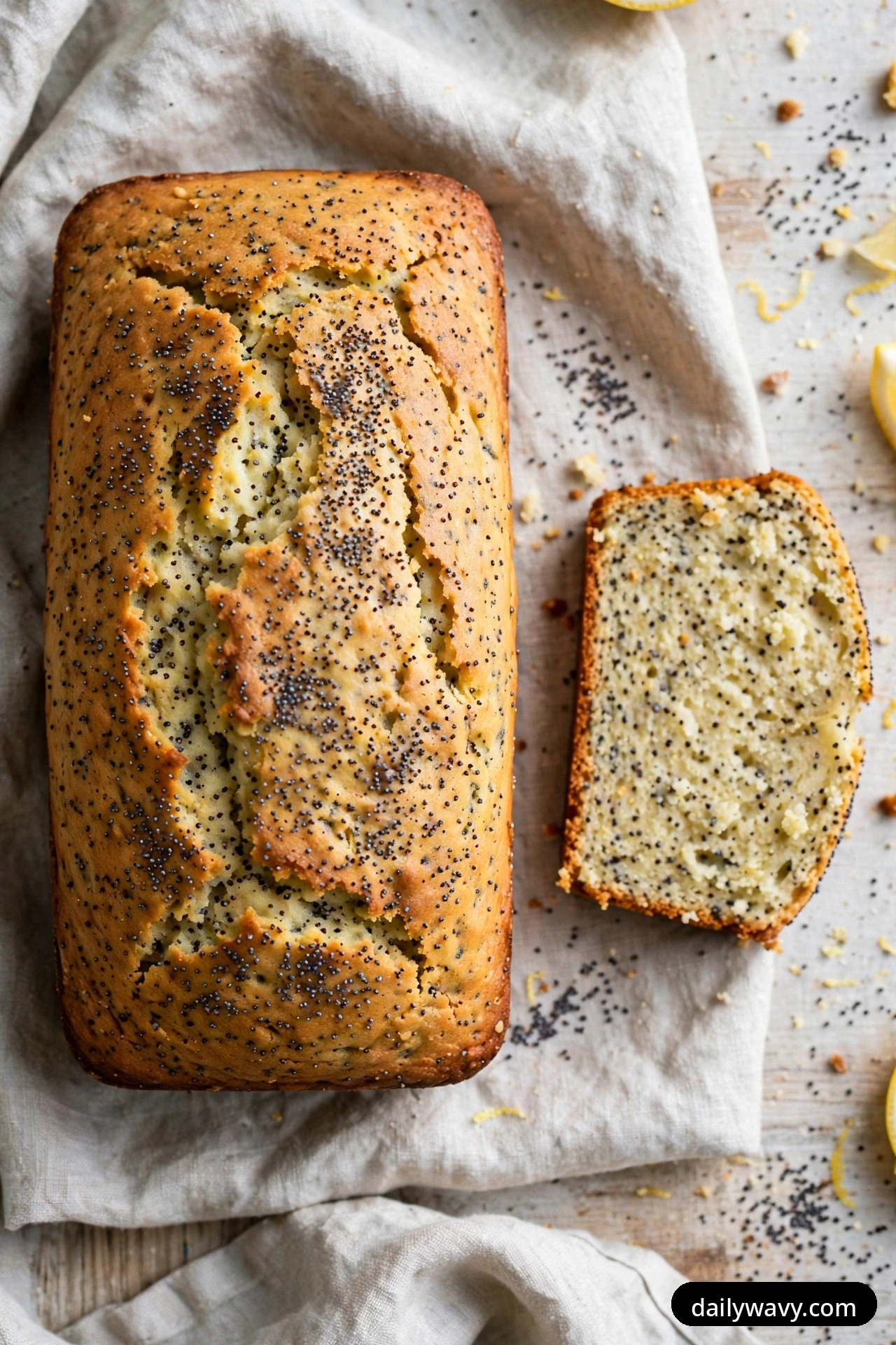 Seen from above, a lemon poppy seed loaf rests on a linen cloth with one slice cut beside it, its pale crumb speckled throughout with black poppy seeds.