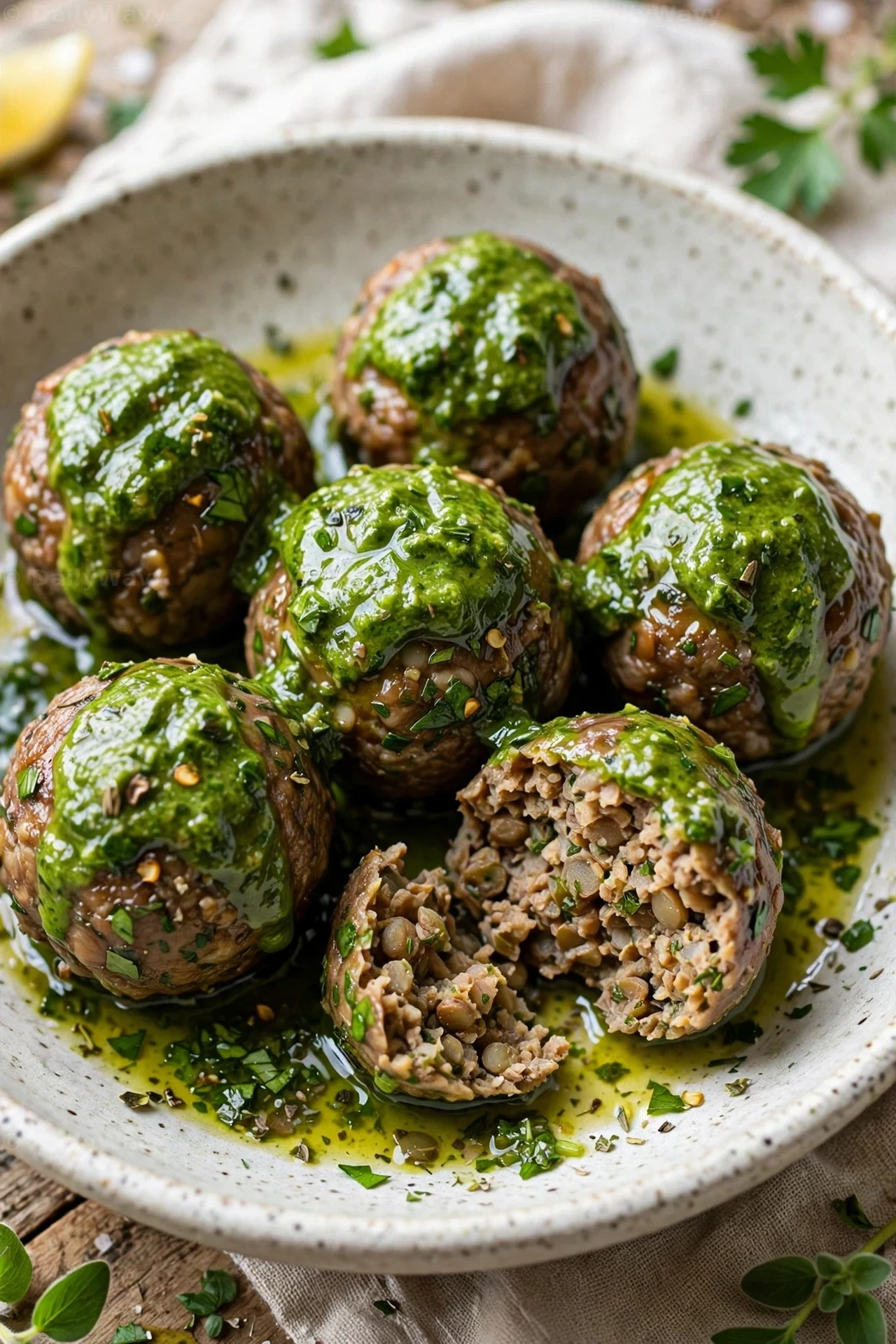 Lentil meatballs spooned with green chimichurri fill a bowl, with one meatball broken open to show the lentil crumb inside.