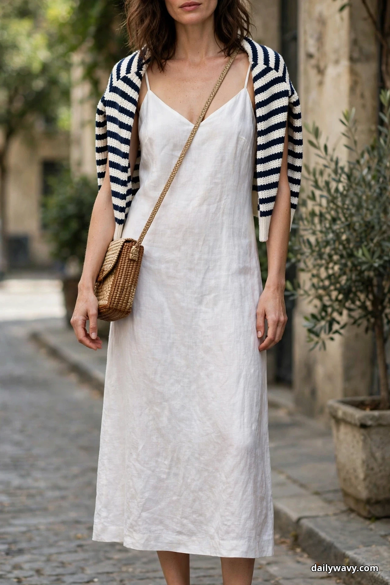 A woman stands on a cobbled street in a frame cropped at the mouth and just below the dress hem, wearing a white linen slip midi dress with a navy-and-white striped knit sweater draped over her shoulders and a woven crossbody bag