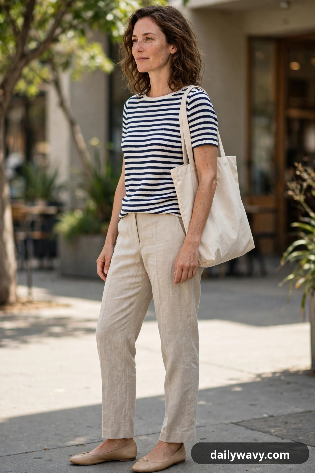 A woman wears a navy and white striped knit top with sand linen trousers, nude ballet flats and a natural canvas tote over her shoulder